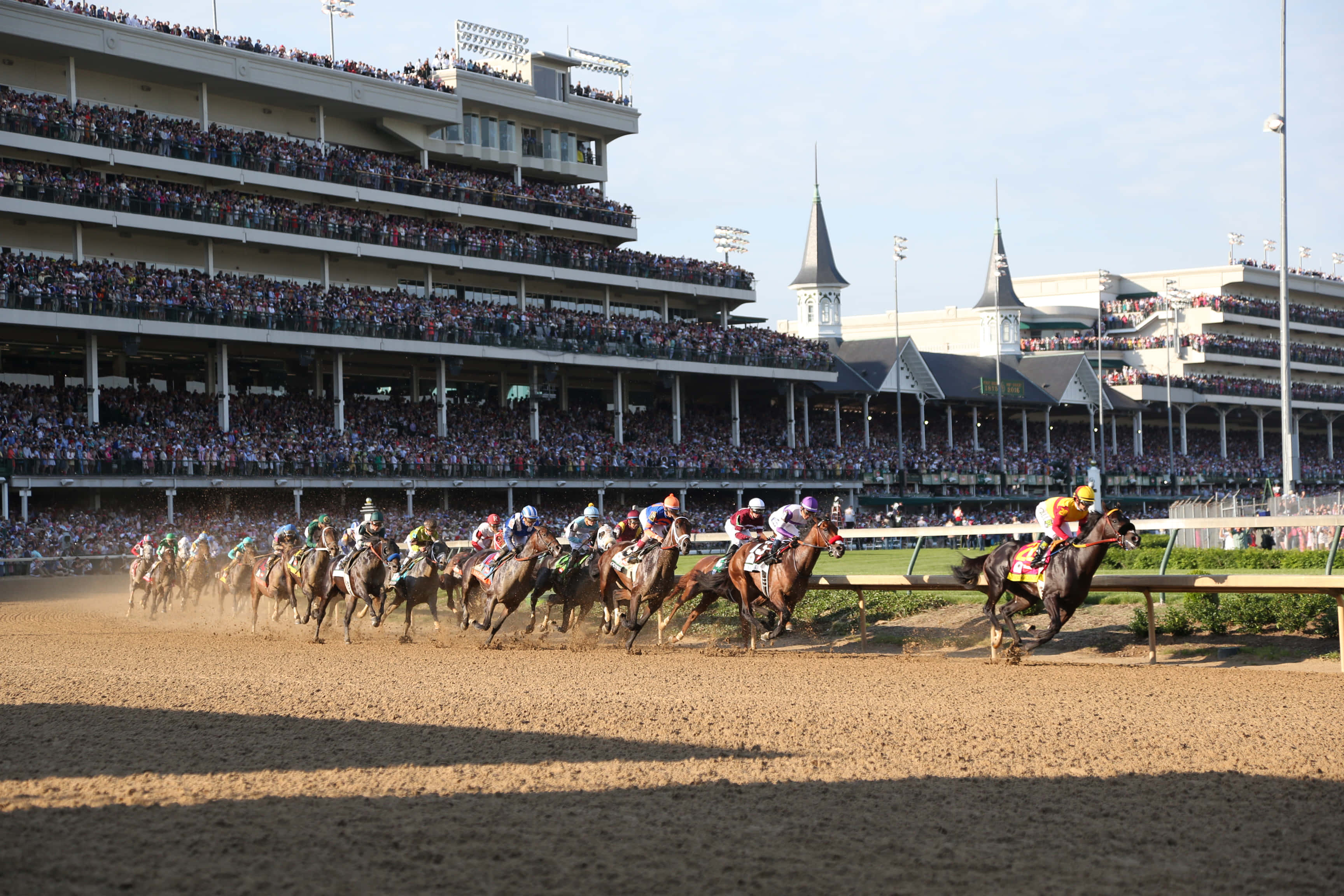 Kentucky Derby 桌布 壁紙 — 騎師在陽光照耀的泥地賽道上策騎純種馬競賽，觀眾在晴朗天空下擠滿看台。