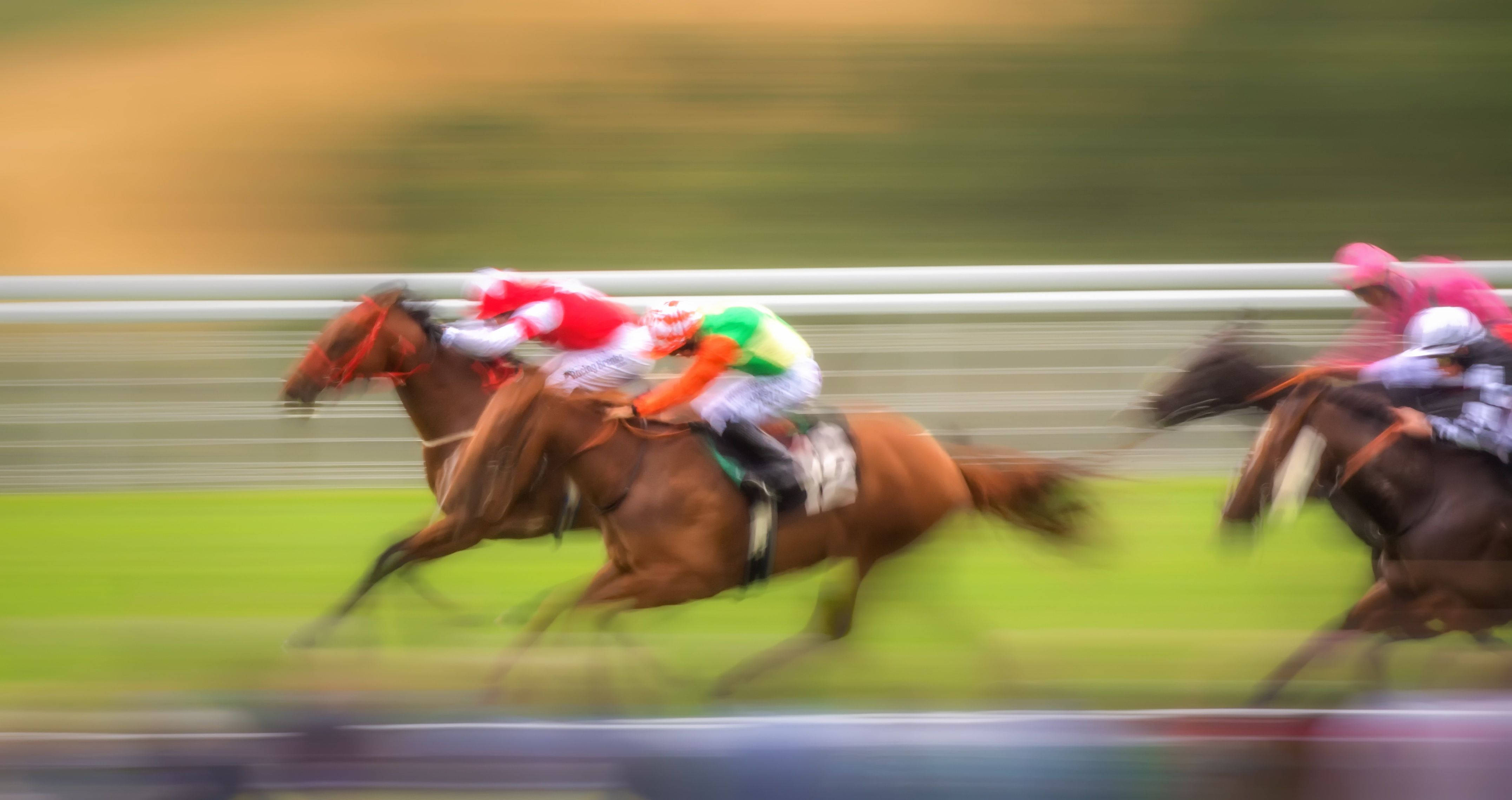 Horse Racing 桌布 壁紙 — 騎師穿著色彩繽紛的絲綢，在高速運動中騎著奔騰的馬匹，穿過模糊的綠色跑道和圍欄。