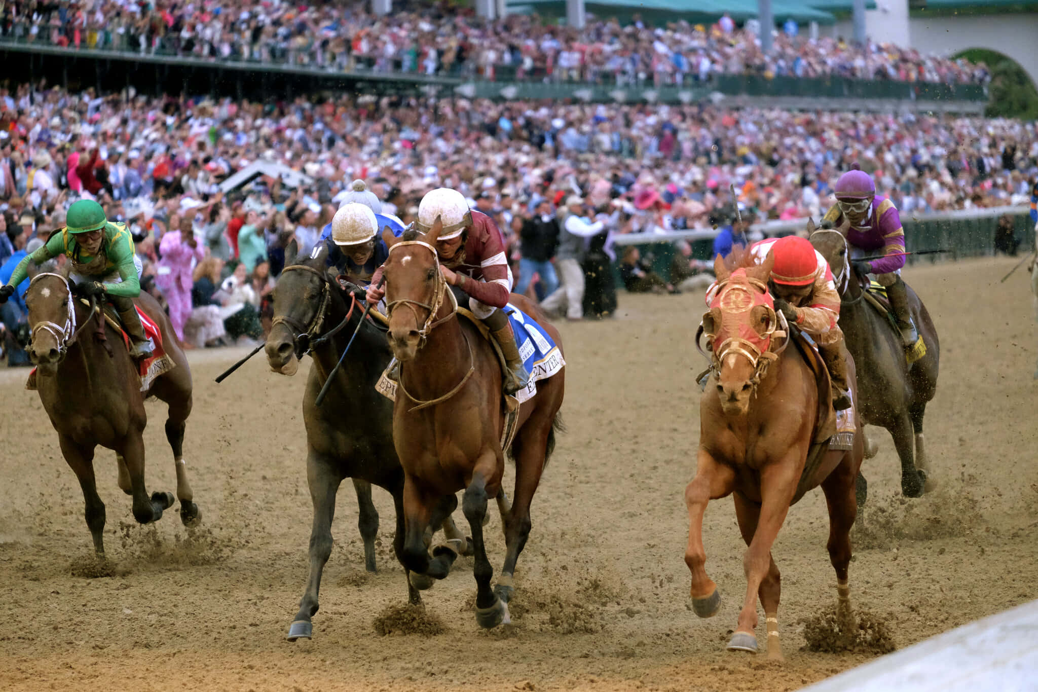 Kentucky Derby 桌布 壁紙 — 騎師穿著色彩繽紛的賽馬服，在滿座的體育館背景下，於泥地賽道上策騎純種馬全速奔馳。