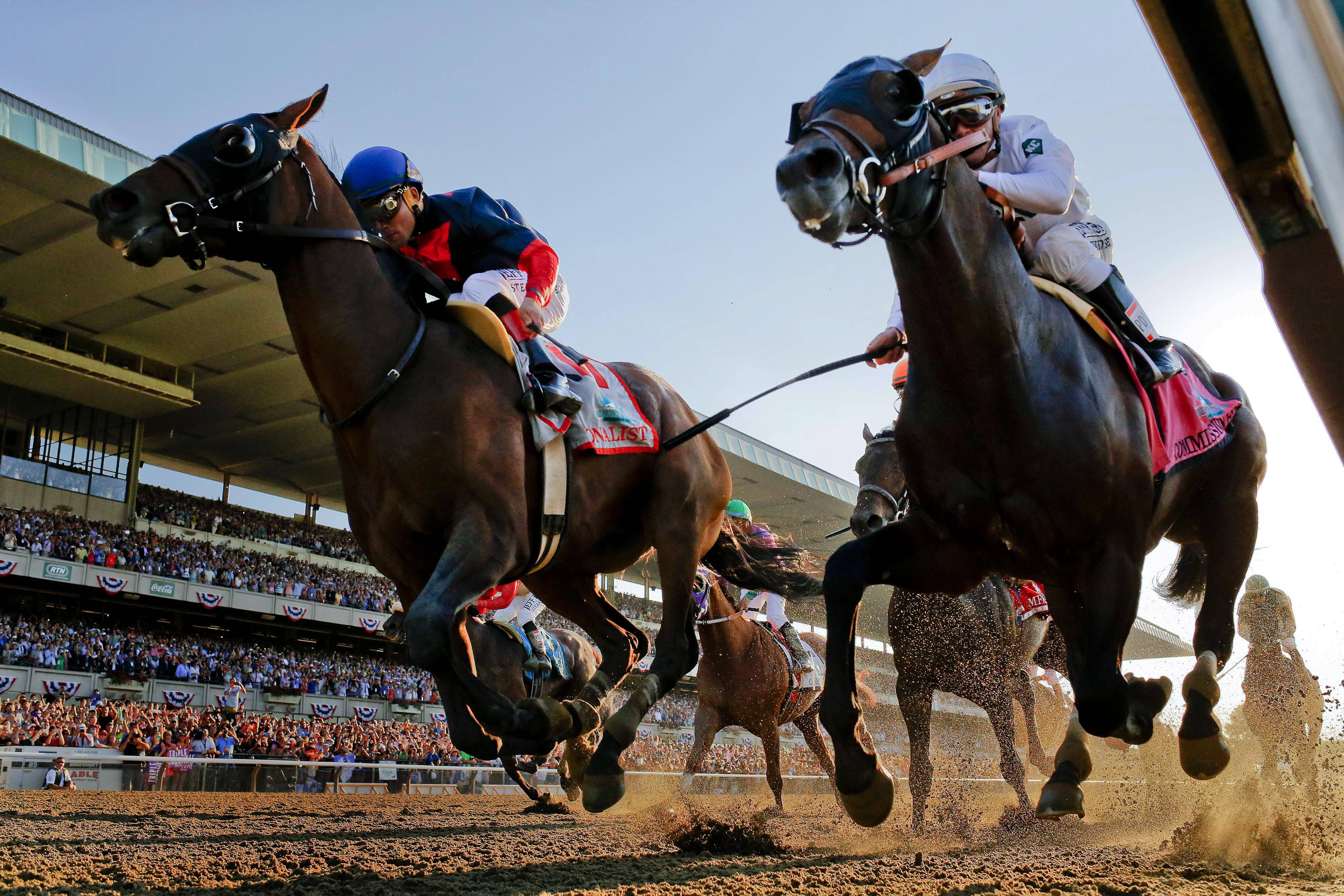 Horse Racing 桌布 壁紙 — 騎師策騎純種馬在泥地賽道上全速奔馳，看台上的人群在明亮的日光下歡呼。