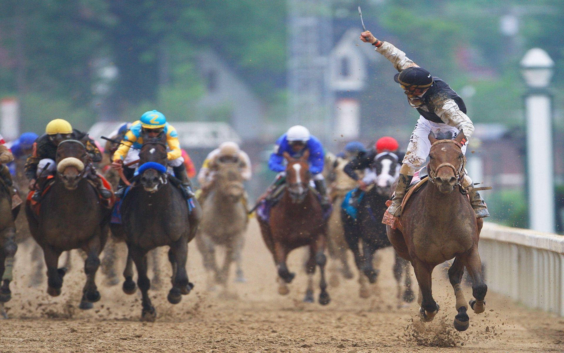 Horse Racing 桌布 壁紙 — 騎師身穿色彩繽紛的絲綢，在泥濘飛濺、背景建築模糊的泥地上策馬奔馳。