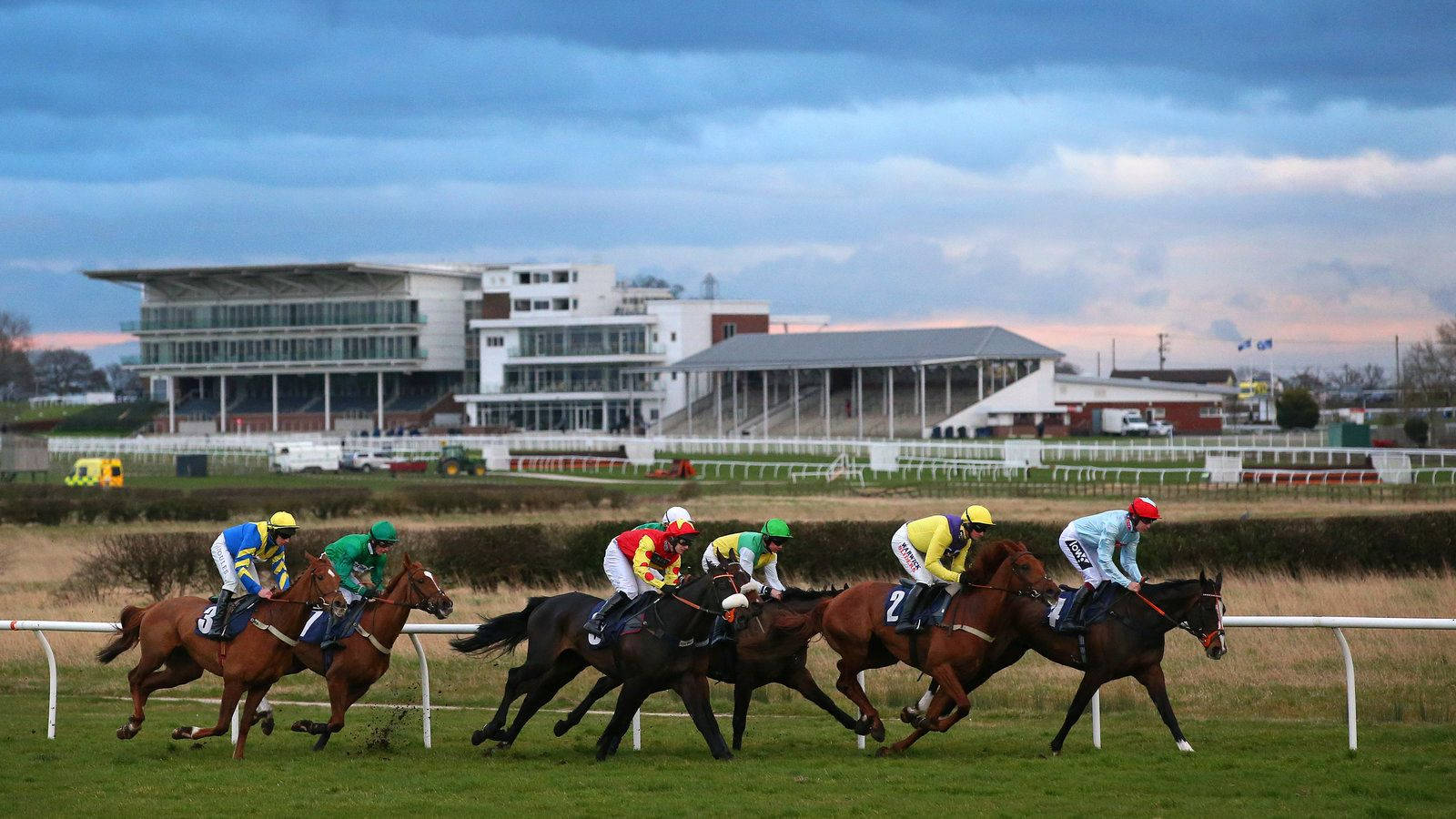 Horse Racing 桌布 壁紙 — 騎師身穿色彩繽紛的綵衣，在草地跑道上策騎純種馬競賽，現代的看台在陰雲密布的暮色天空下若隱若現。