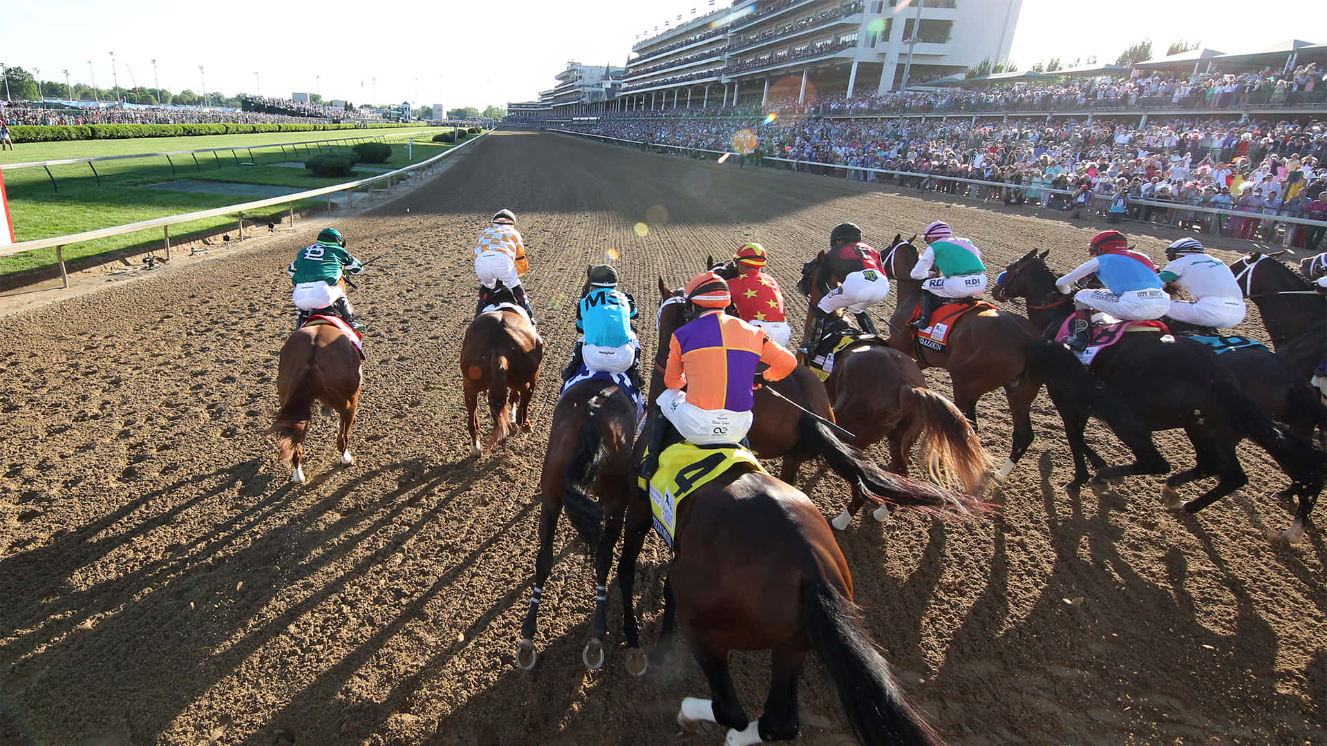 Kentucky Derby 桌布 壁紙 — 騎師身穿色彩繽紛的賽馬服，在陽光普照的泥地賽道上策騎純種馬競賽，觀眾在看台上歡呼。