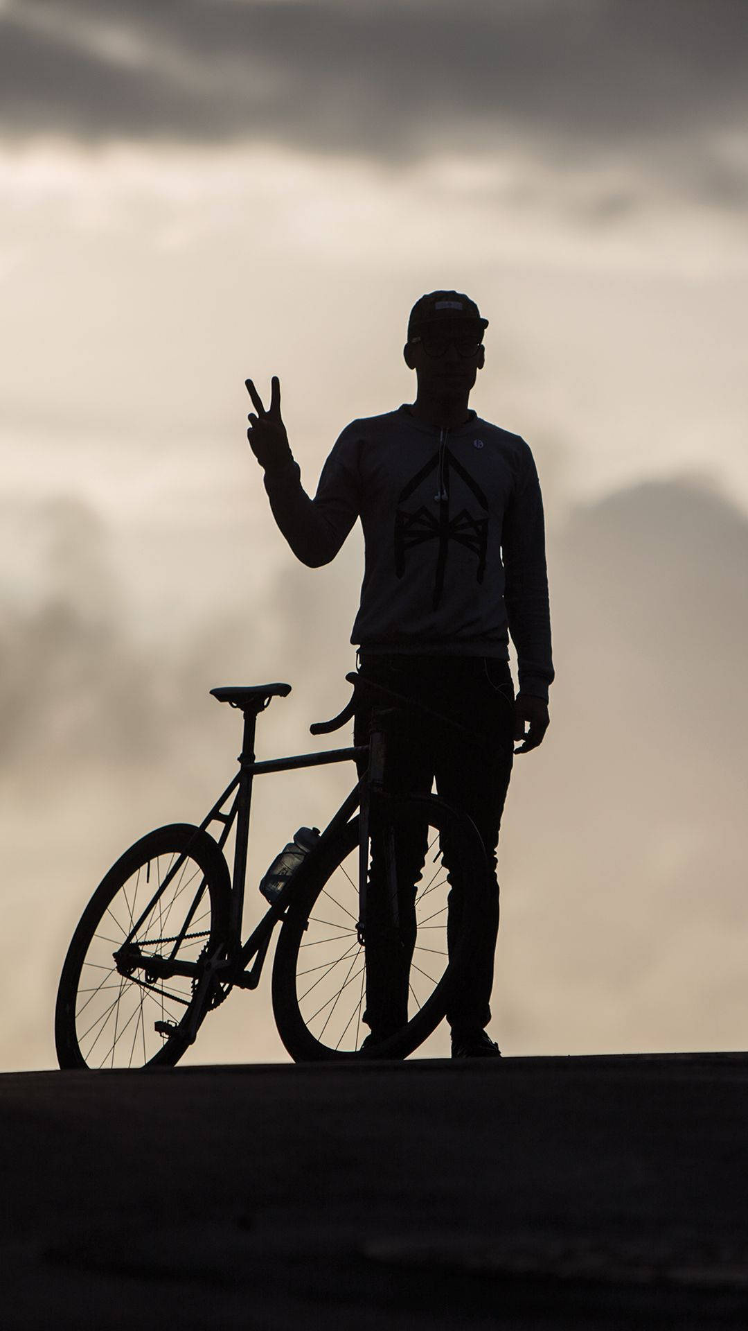 Silhouette 桌布 壁紙 — 騎自行車者的剪影站在自行車旁，在陰雲密佈的天空下做出和平手勢。