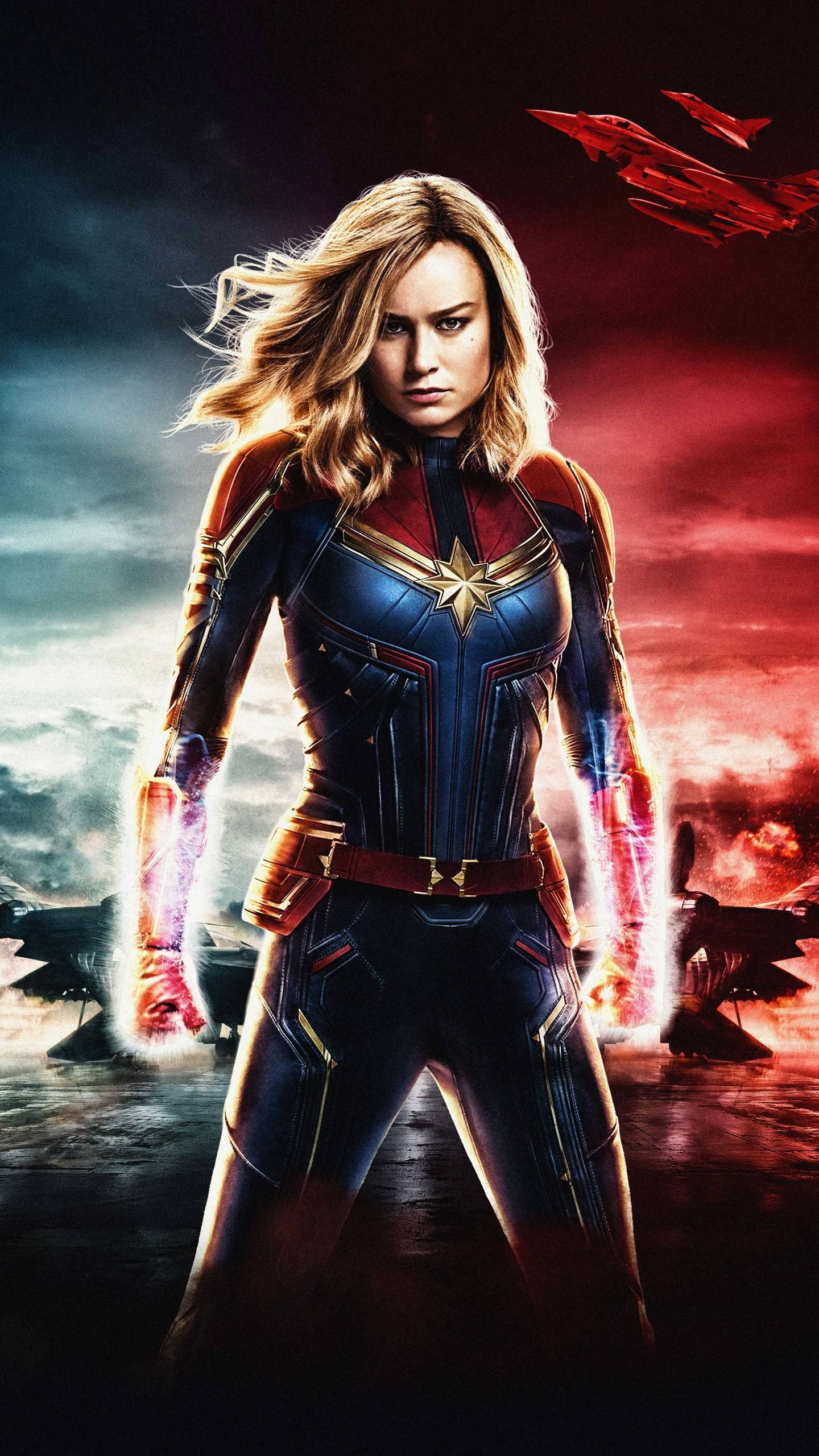 captain marvel iPhone 桌布 壁紙 — 驚奇隊長以充滿力量的動態姿勢站立，發光的能量拳頭對比著戲劇性的紅色和灰色天空，戰鬥機在旁。