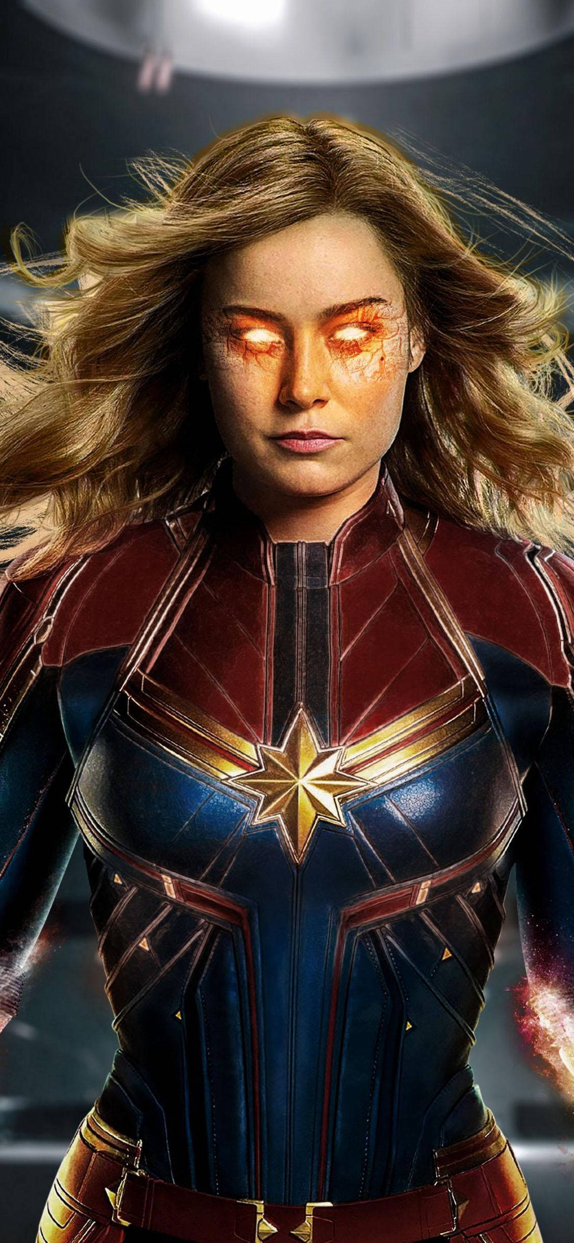 captain marvel iPhone 桌布 壁紙 — 驚奇隊長以動感姿勢站立，發光的橙色眼睛和飄逸的金色頭髮，背景是黑暗的科幻風格。