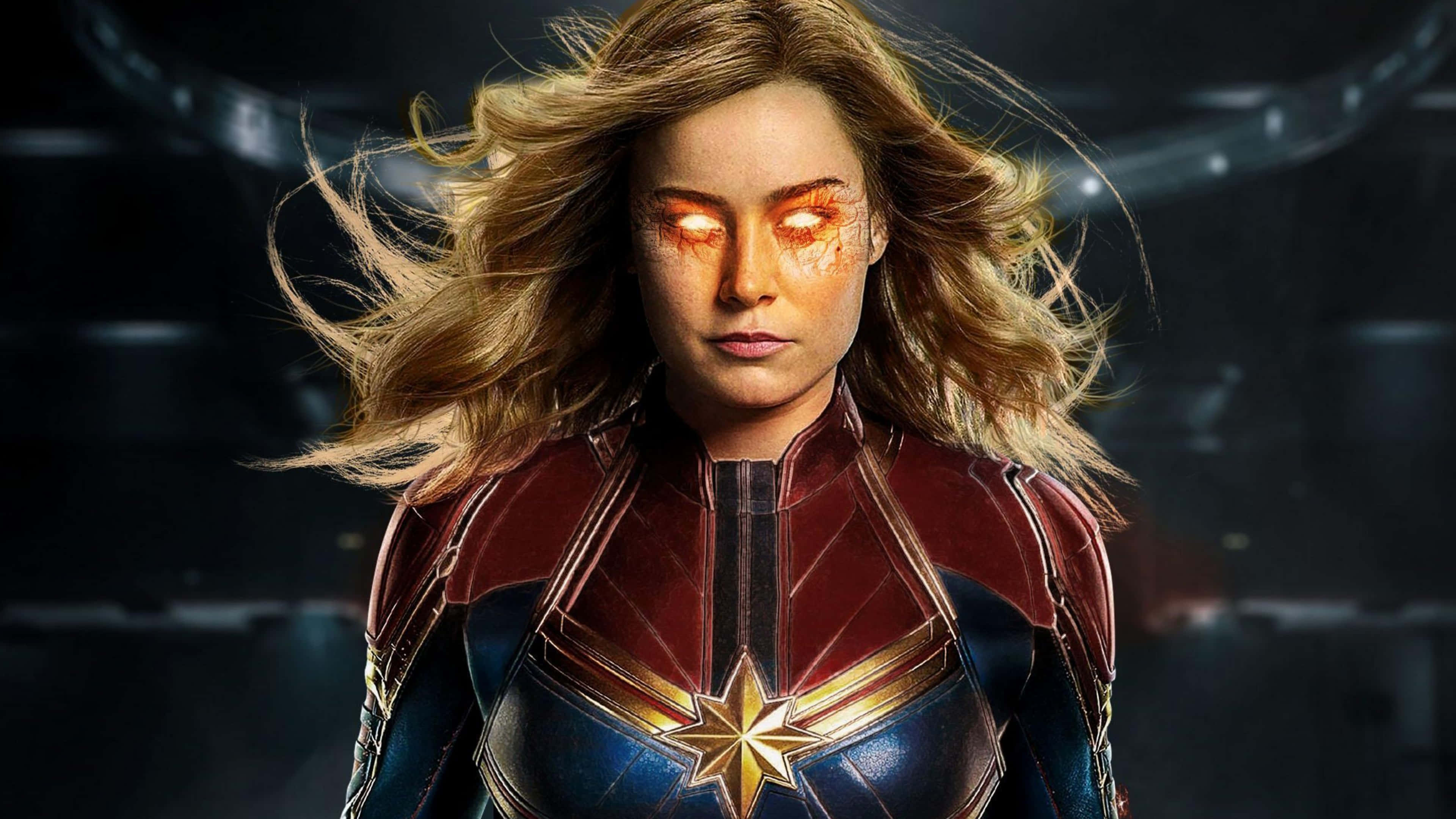 4K Marvel 壁紙 桌布 壁紙 — 驚奇隊長在黑暗科幻背景下，身穿紅藍戰衣，散發著橙色光芒的眼睛和飄逸的金色頭髮。