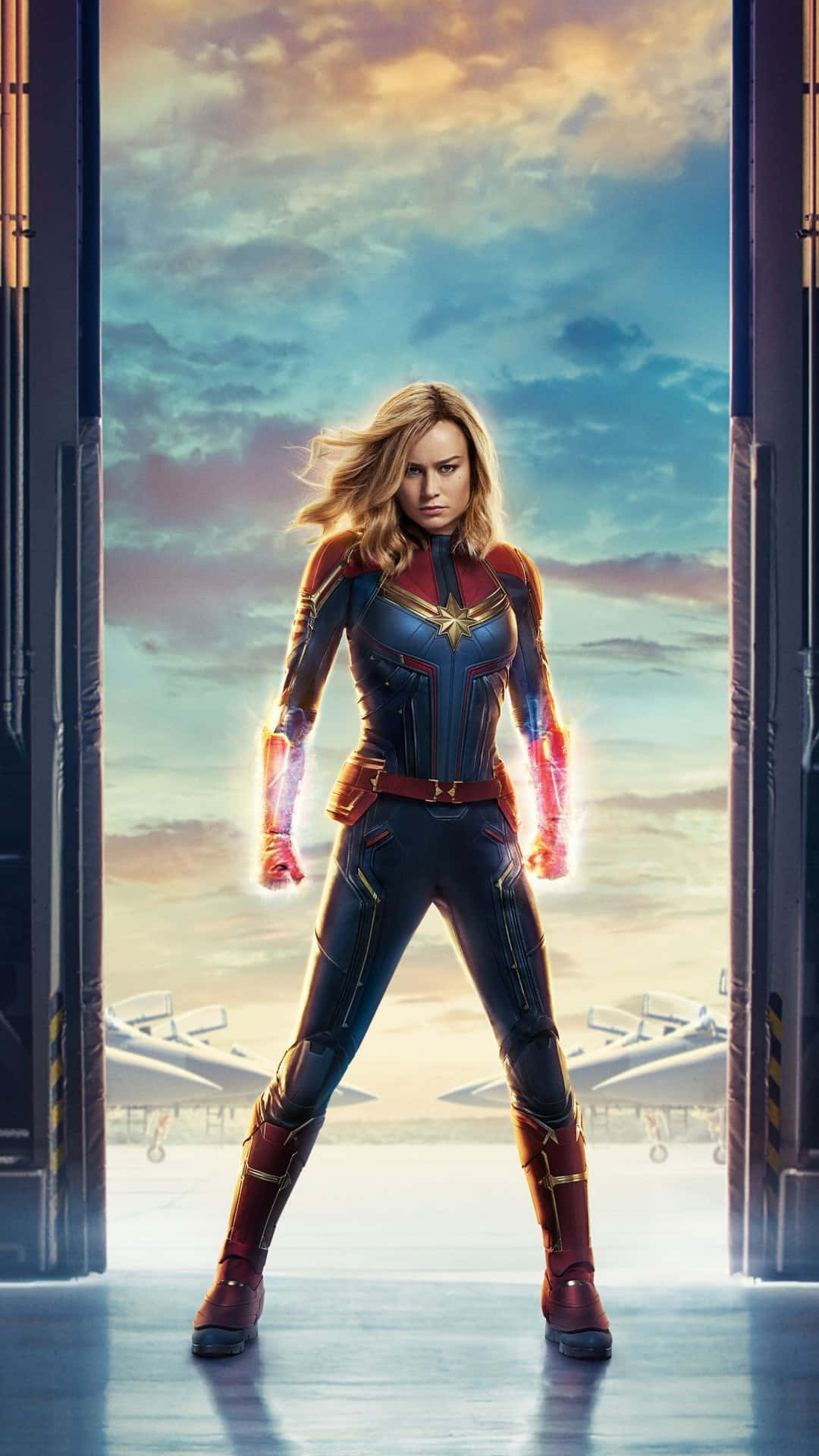 captain marvel iPhone 桌布 壁紙 — 驚奇隊長強勢地站在機庫門口，拳頭發光，背景是充滿活力的日落天空。