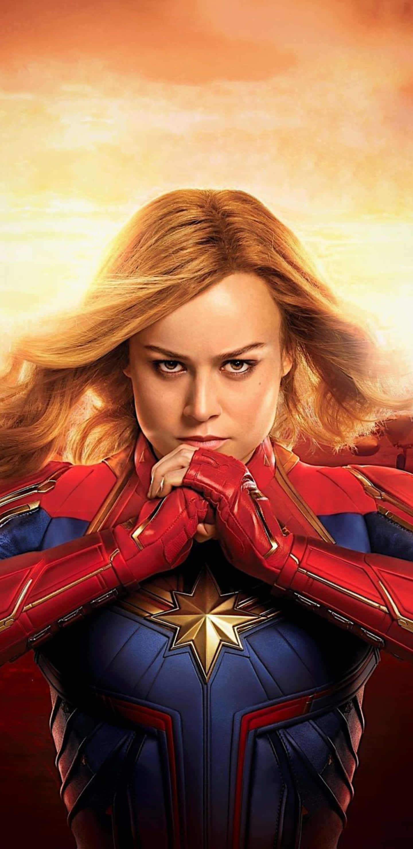 Pixel 3xl Captain Marvel 桌布 壁紙 — 驚奇隊長自信地穿著她的紅藍戰衣，站在發光的橙色天空前，風吹動她的金髮。