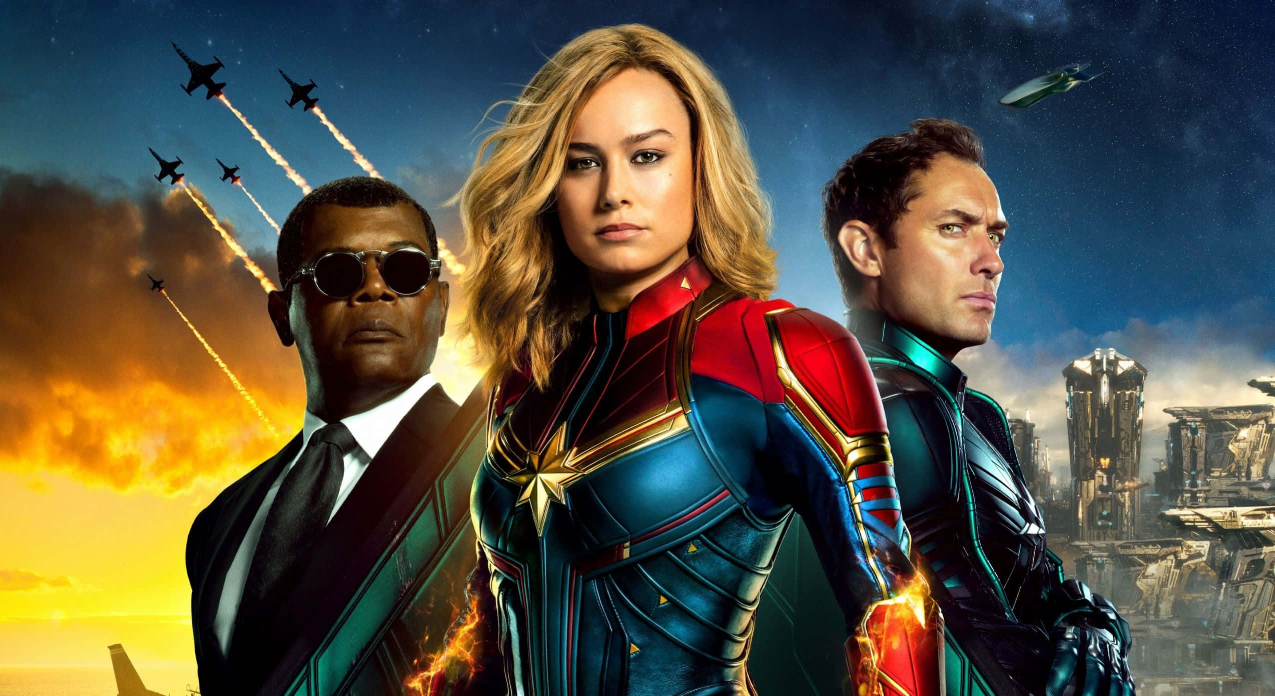 Captain Marvel 桌布 壁紙 — 驚奇隊長與尼克·福瑞和獵鷹英勇地站在熾熱的日落天空下，背景有戰鬥機和未來主義城市景觀。