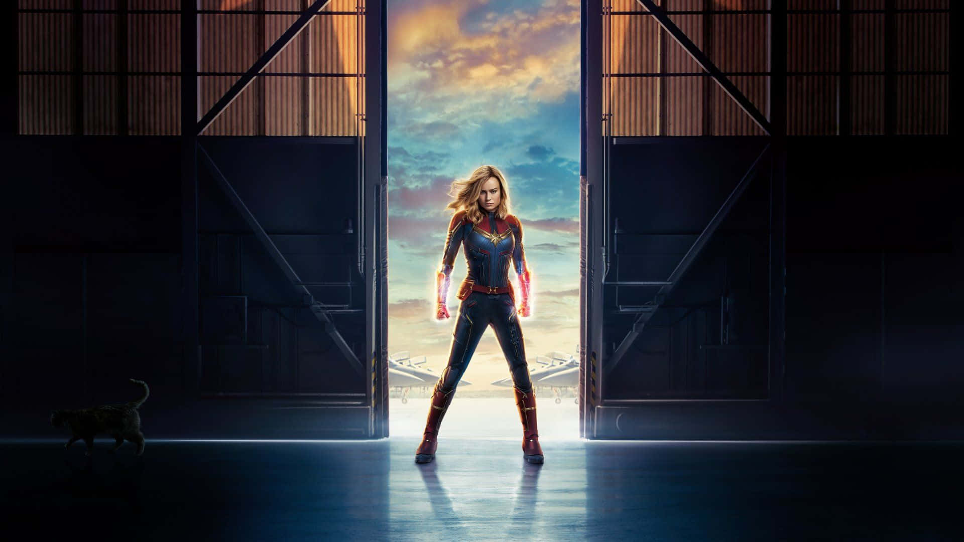 Captain Marvel HD 桌布 壁紙 — 驚奇隊長英勇地站在機庫門口，金色光芒從她身後湧入，照亮她發光的拳頭，一隻貓在陰影中走過。