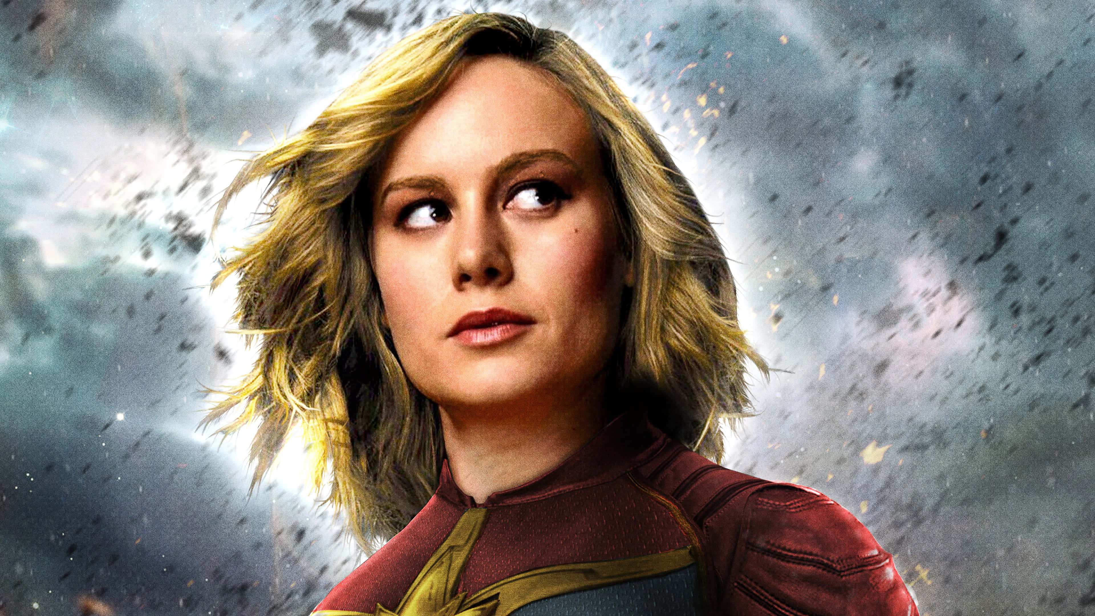 brie larson 桌布 壁紙 — 驚奇隊長身穿紅藍戰衣，在宇宙碎片和發光粒子的風暴中向上凝視。