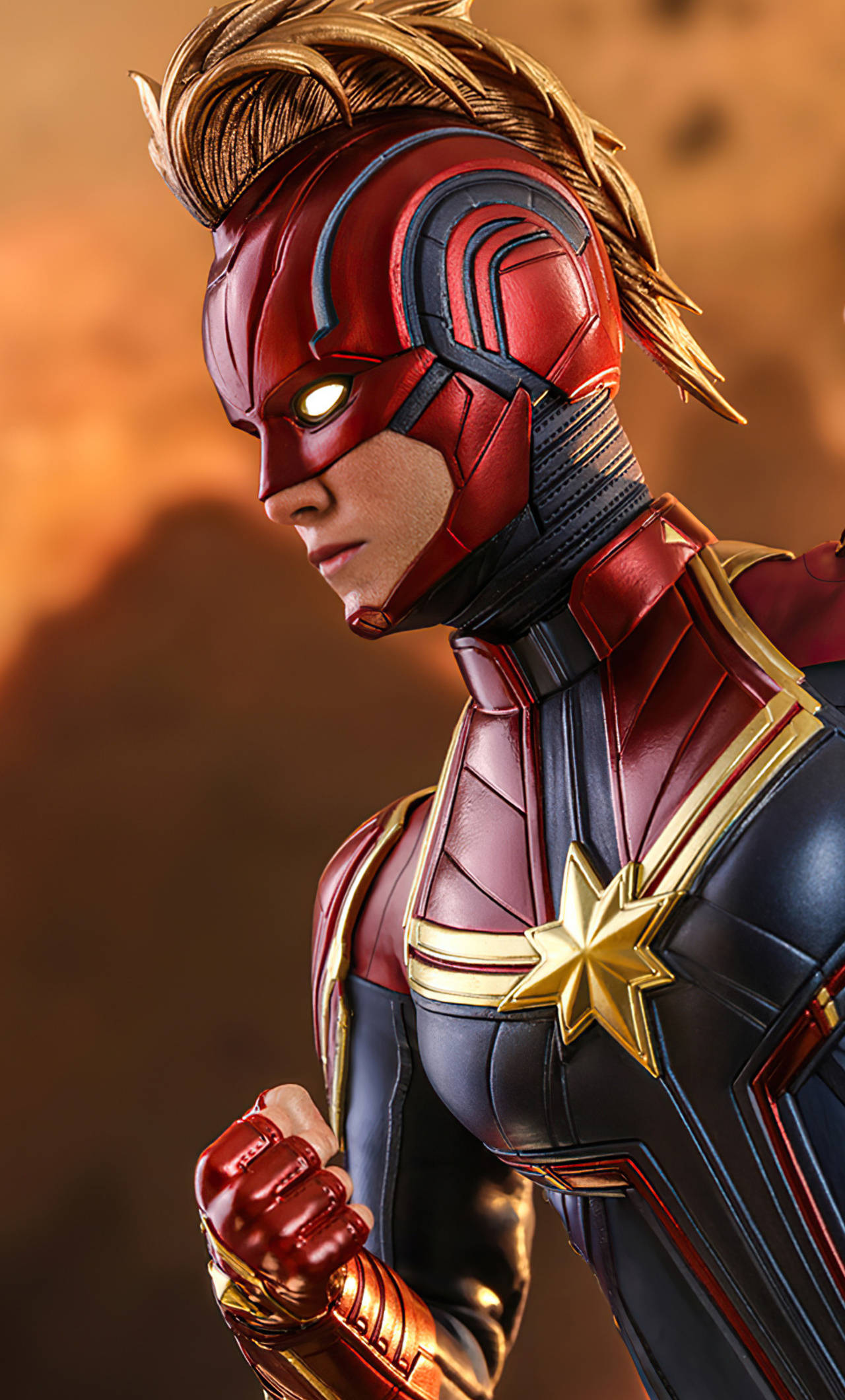 captain marvel iPhone 桌布 壁紙 — 驚奇隊長身穿紅金藍色戰衣，發光眼睛，握緊拳頭，背景為溫暖的橙色。