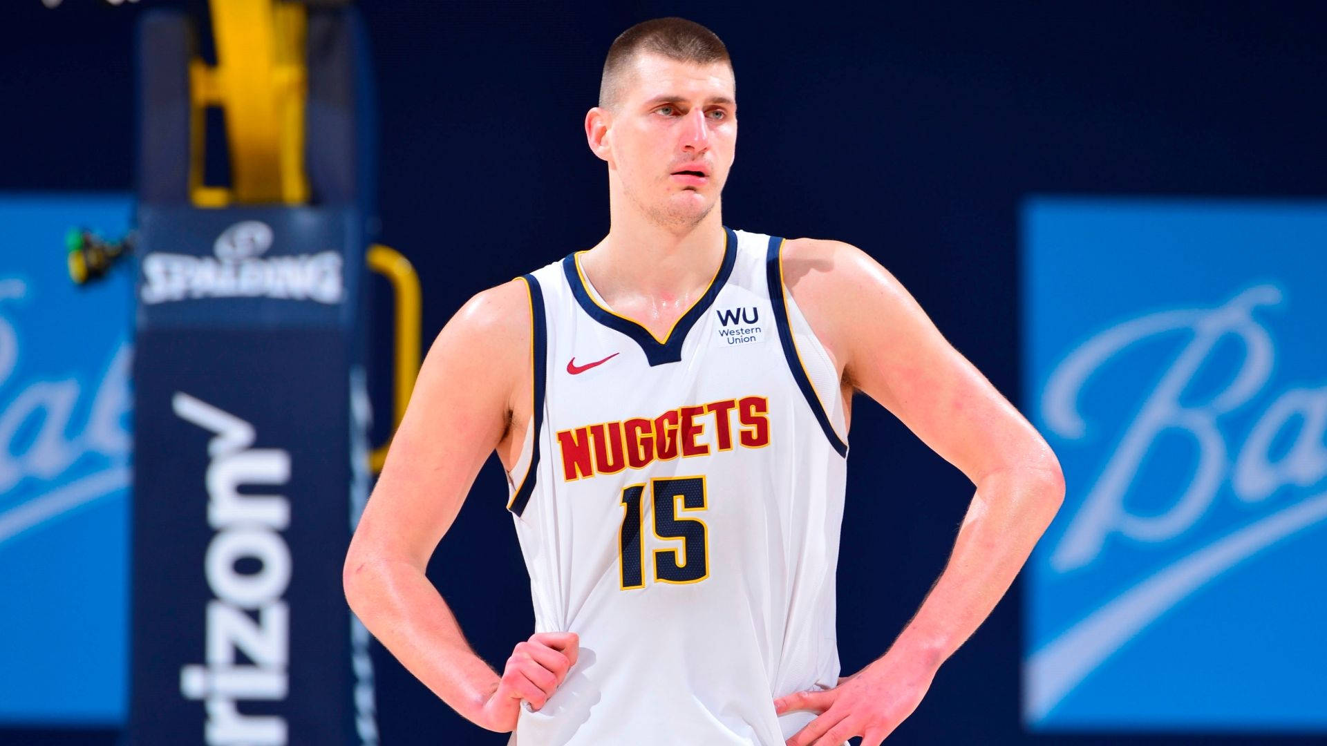 Jokic 桌布 壁紙 — 高大的籃球運動員身穿白色丹佛金塊隊15號球衣，在明亮的場館燈光下雙手叉腰站在球場上。