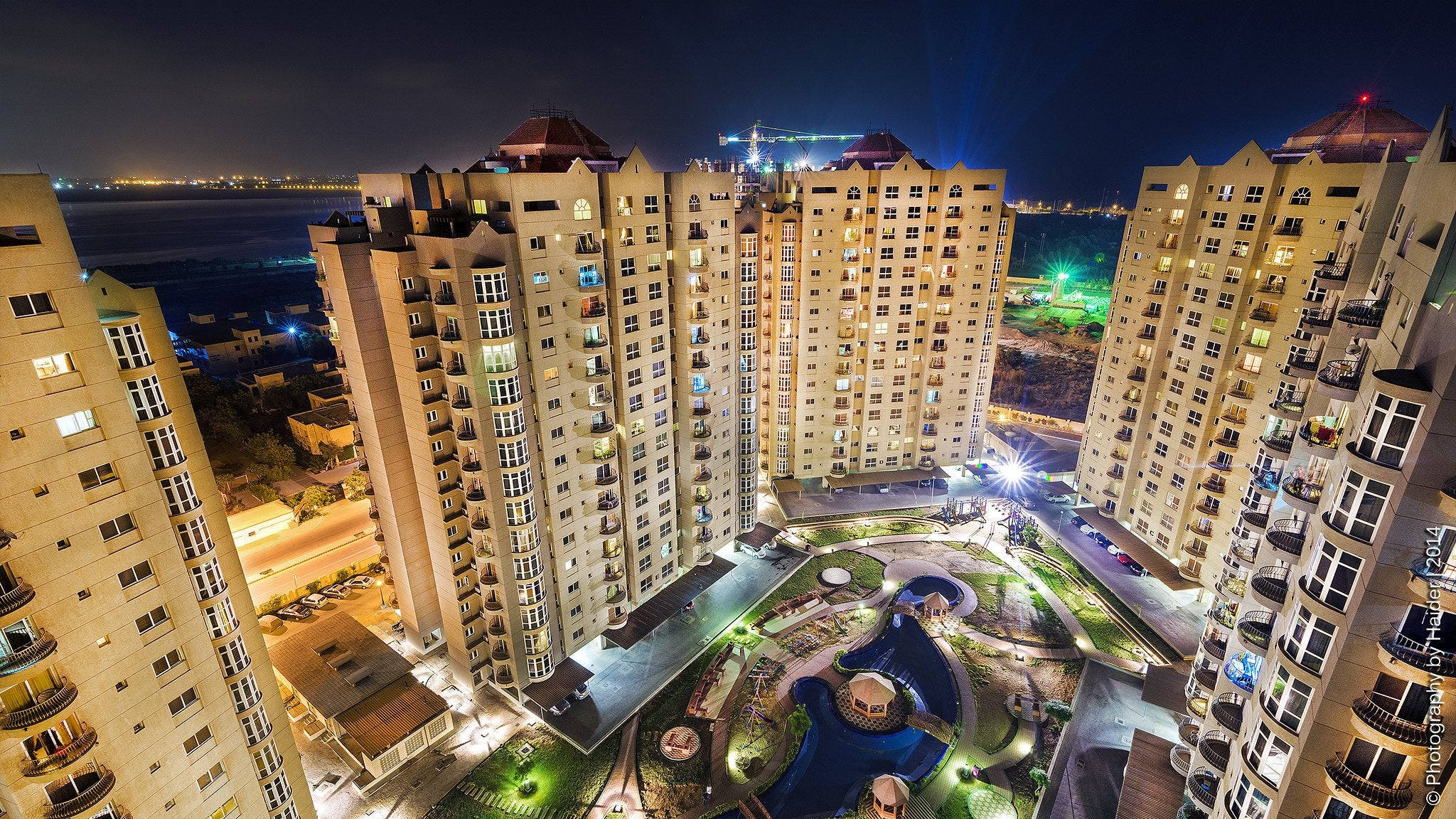 Karachi 桌布 壁紙 — 高層住宅公寓的夜間空中景觀，燈火通明，環繞著一個帶有游泳池和小徑的園景庭院。