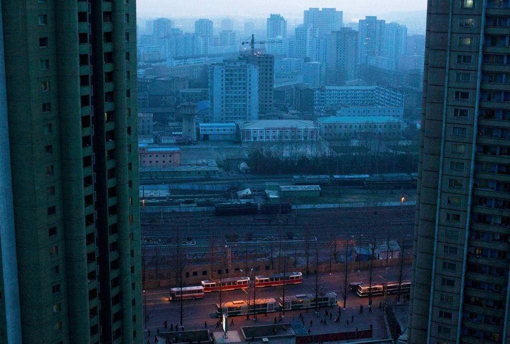 韓國 桌布 壁紙 — 高樓大廈框住的朦朧城市景觀，顯示電車在黃昏時沿著軌道行駛，帶有柔和的藍色和灰色色調。