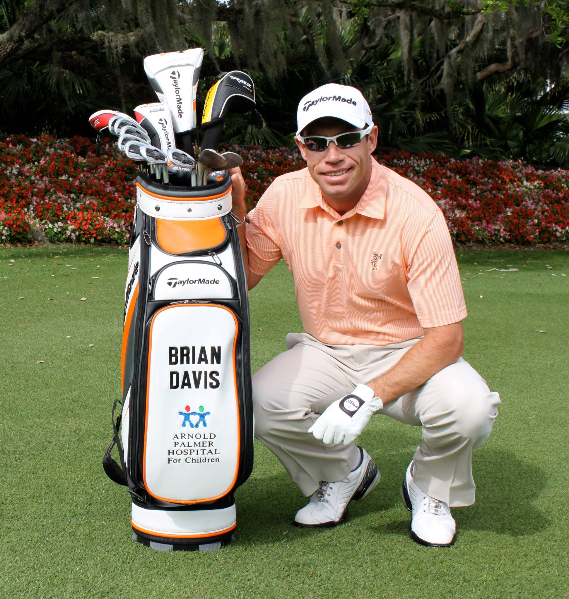 高爾夫 桌布 壁紙 — 高爾夫球手 Brian Davis 跪在他 TaylorMade 球袋旁，身後是茂盛的綠色球場和開花的灌木叢。