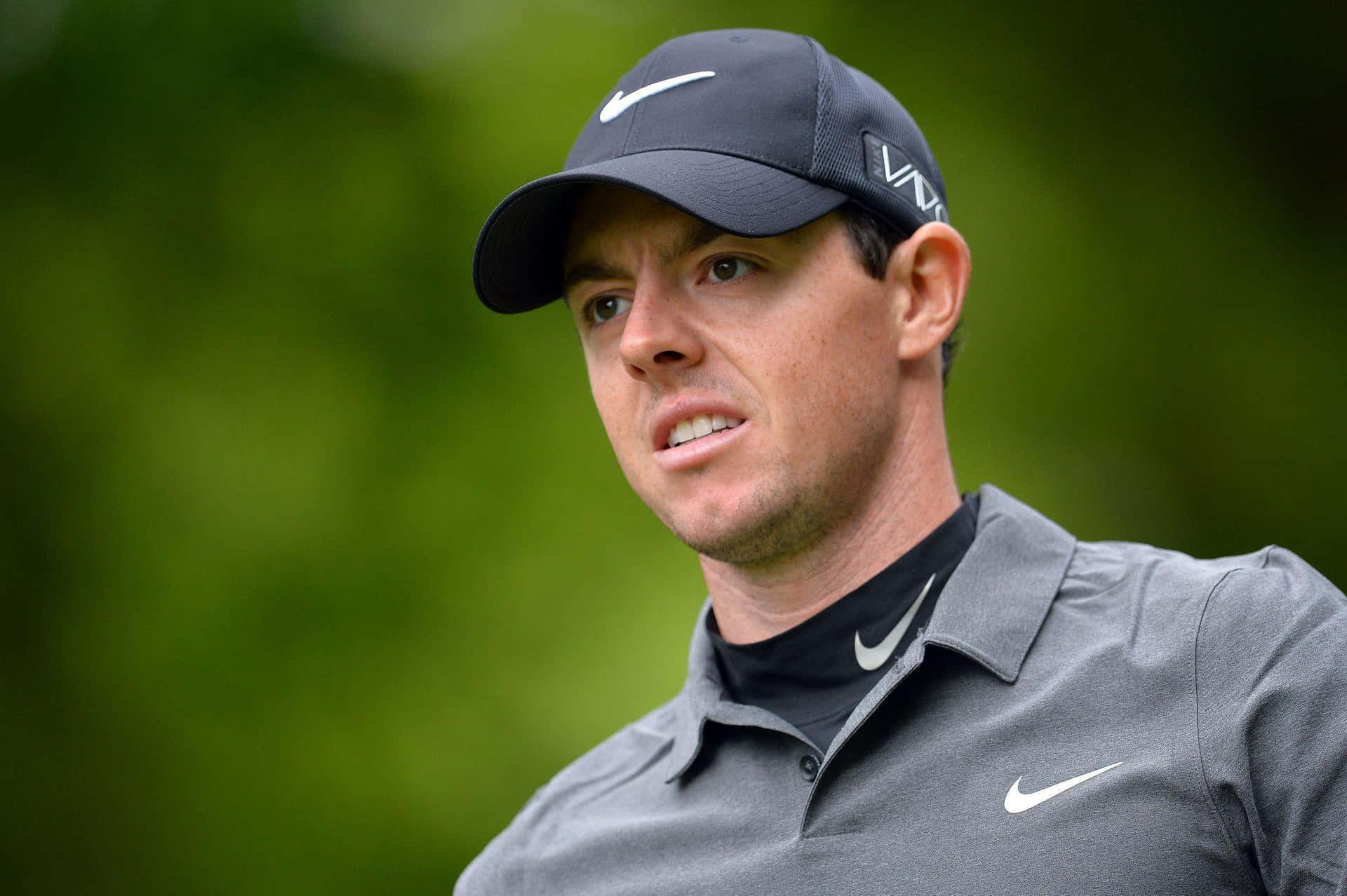  壁紙 — 高爾夫球手Rory McIlroy專注的表情，戴著深色帽子，穿著灰色Nike Polo衫，背景是模糊的綠色。