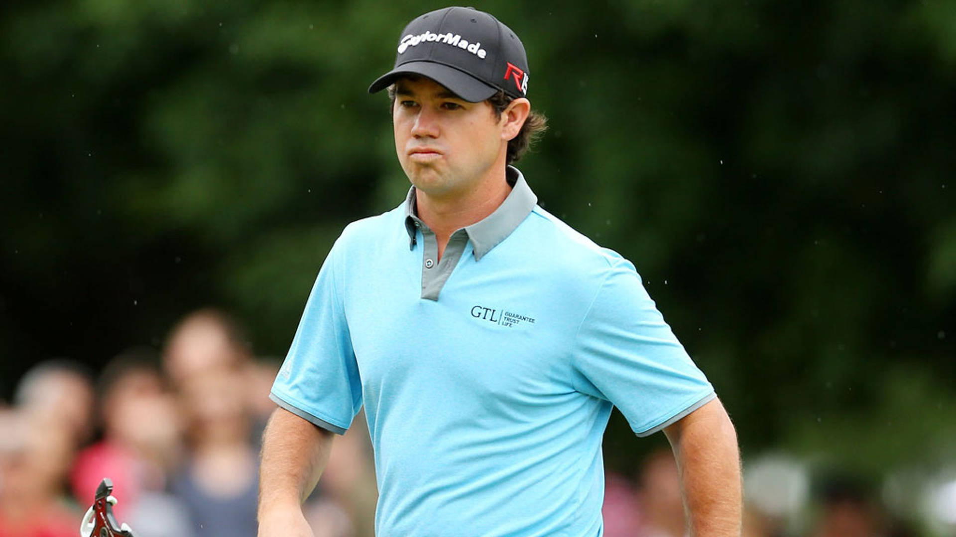  壁紙 — 高爾夫球手Rory McIlroy身穿淺藍色Polo衫和黑色帽子，專注地站在球場上，背景是模糊的觀眾。