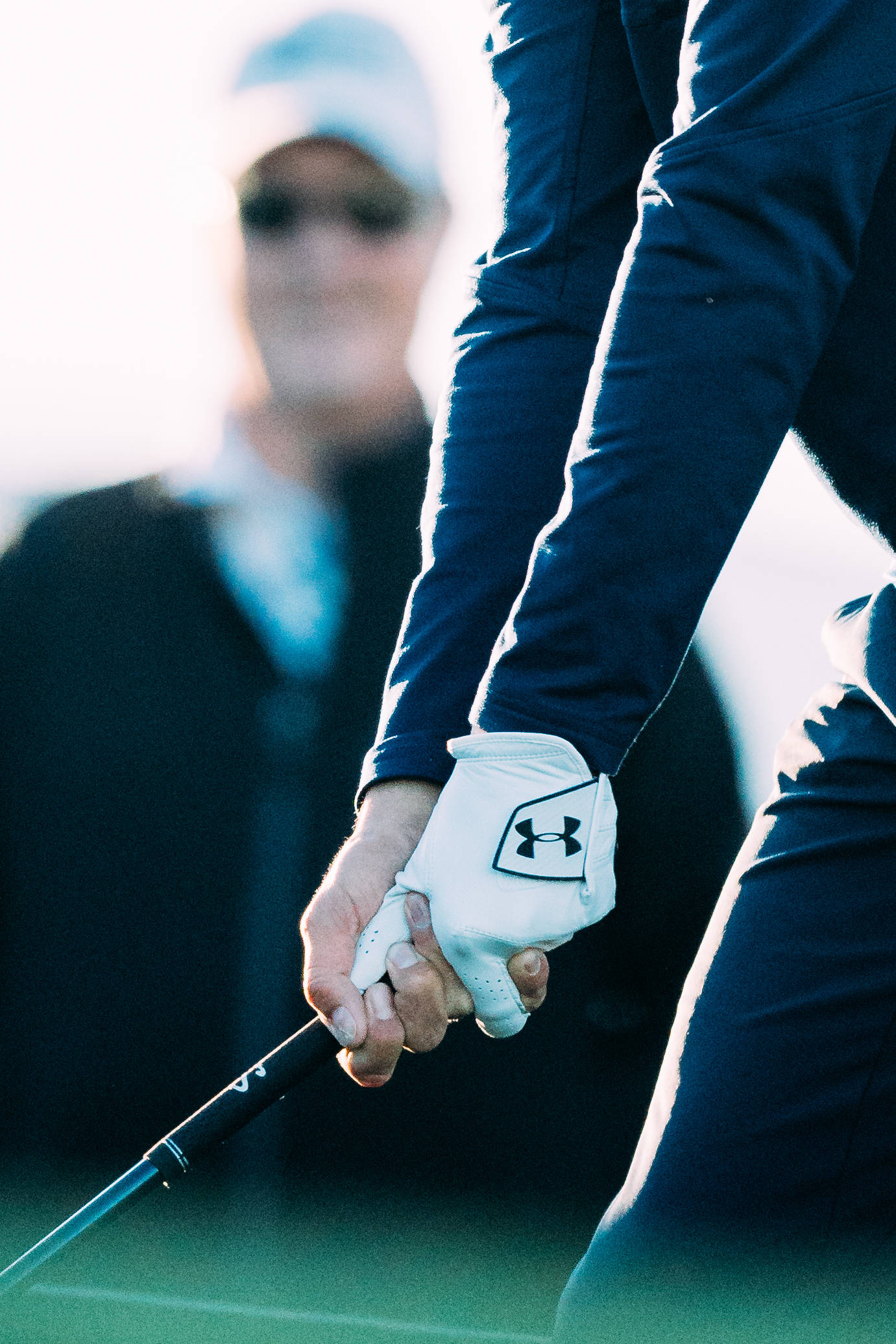 Under Armour 桌布 壁紙 — 高爾夫球手戴手套的手緊握球桿的特寫，背景有模糊的桿弟，在明亮的陽光下。