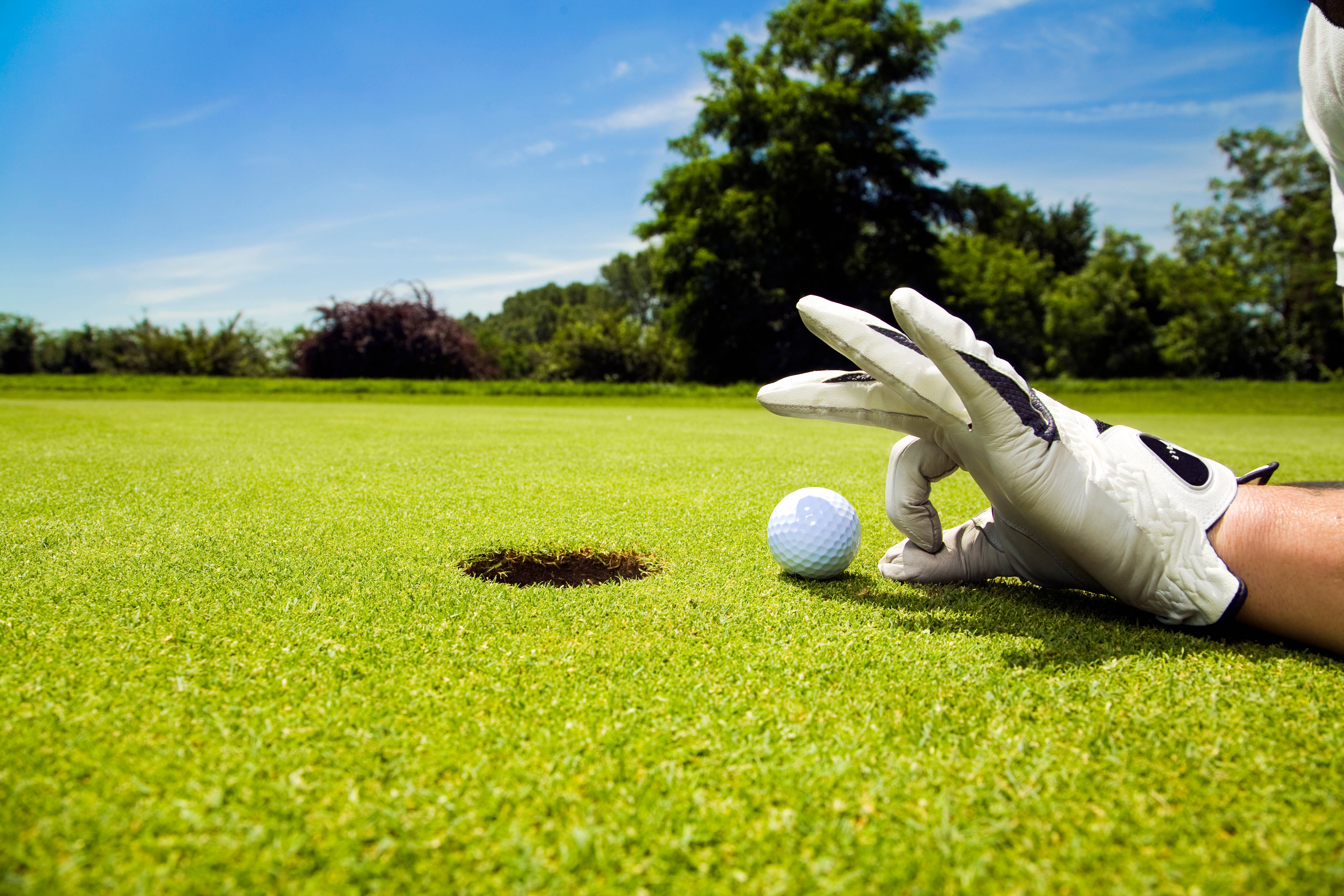 Golfing 桌布 壁紙 — 高爾夫球手戴著手套的手懸停在一個距離球洞僅幾英寸的高爾夫球上方，球位於充滿活力的綠色球道上，背景是晴朗的藍天。
