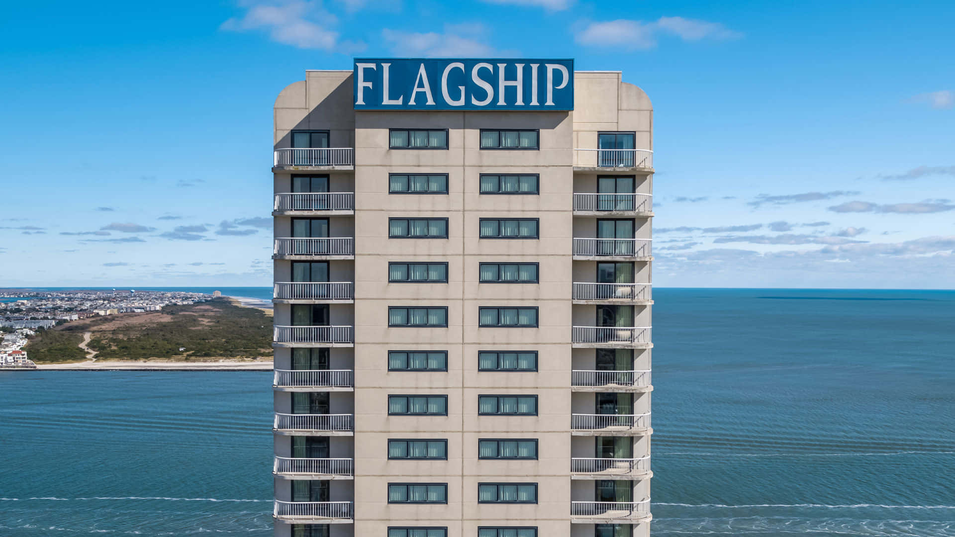 飯店 桌布 壁紙 — 高的米色酒店塔樓帶有'FLAGSHIP'標誌，俯瞰晴朗天空下平靜的藍色海洋。