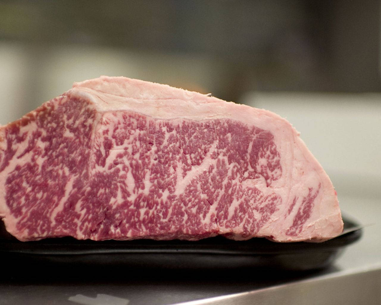 meat 桌布 壁紙 — 高級大理石紋牛肉排特寫，放在黑色盤子上，背景是模糊的廚房。