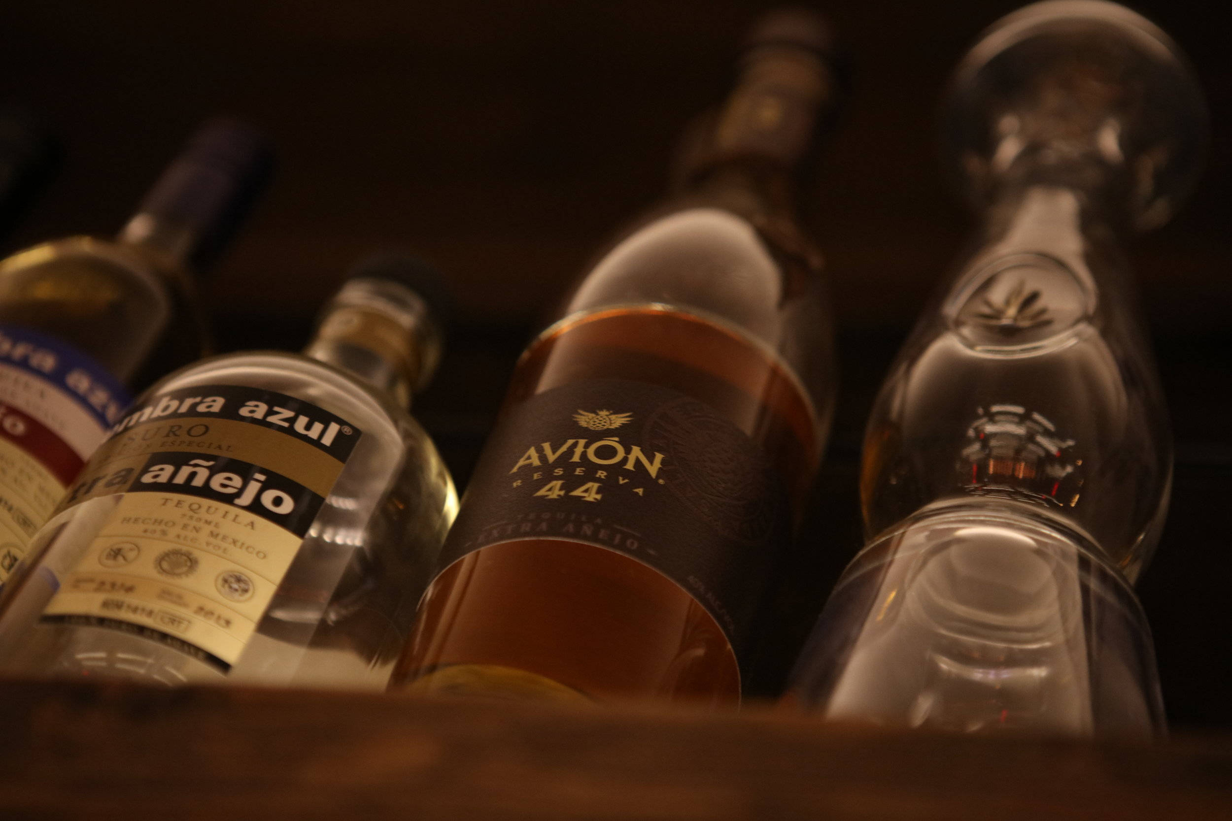 Drink 桌布 壁紙 — 高級龍舌蘭酒瓶和玻璃器皿的特寫，放置在昏暗的木架上，配有溫暖的環境照明。