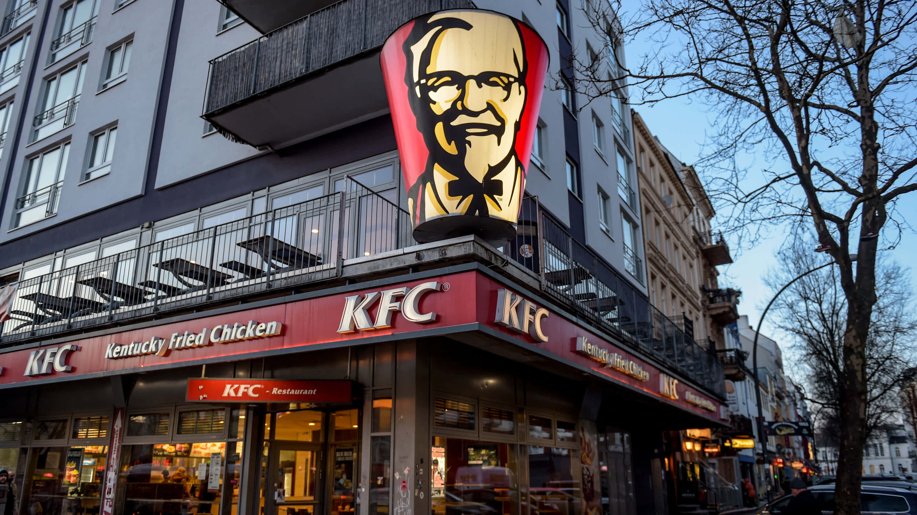 肯德基 桌布 壁紙 — 高聳的KFC桑德斯上校招牌在繁忙的城市餐廳上方發光，帶有紅色品牌和玻璃窗。