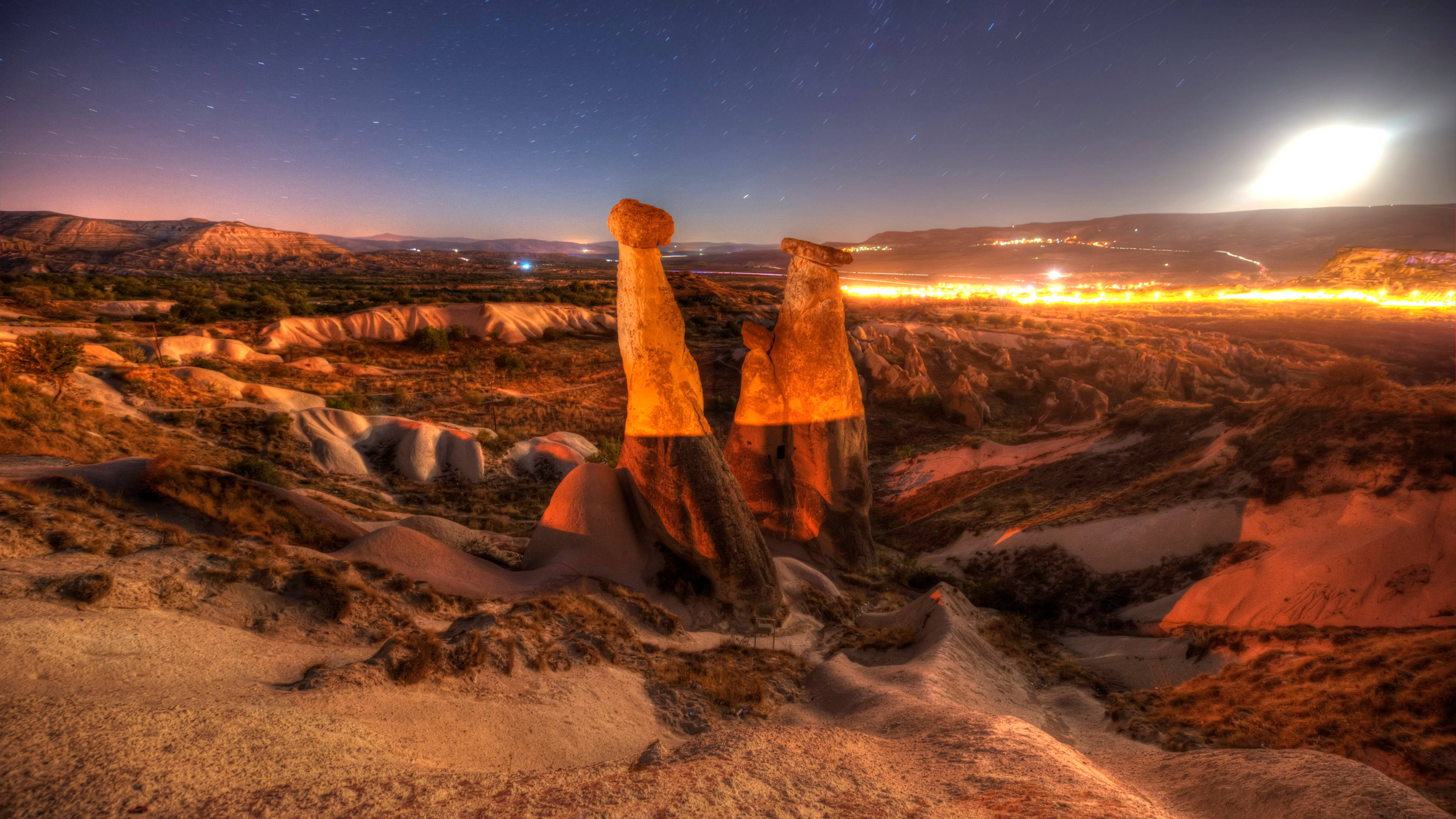 Cappadocia 桌布 壁紙 — 高聳的仙女煙囪在繁星點點的暮色天空下閃耀，伴隨著明亮的月亮和遠處的城市燈光。