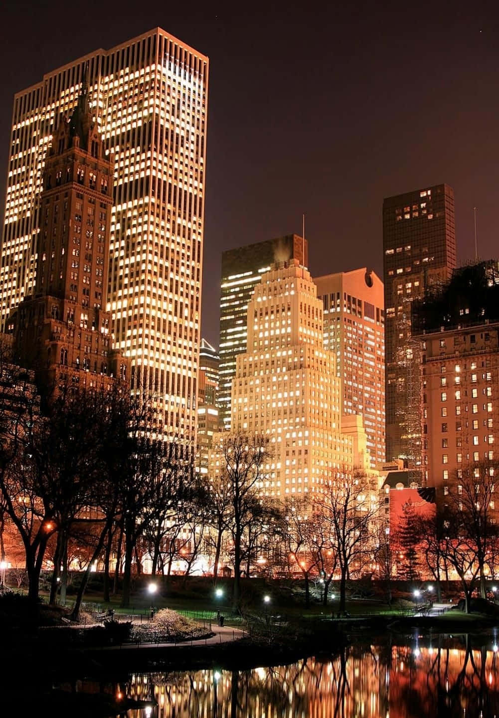 Nyc Aesthetic 桌布 壁紙 — 高聳的城市摩天大樓在夜晚溫暖地發光，它們的燈光倒映在一個被光禿樹木環繞的寧靜池塘中。
