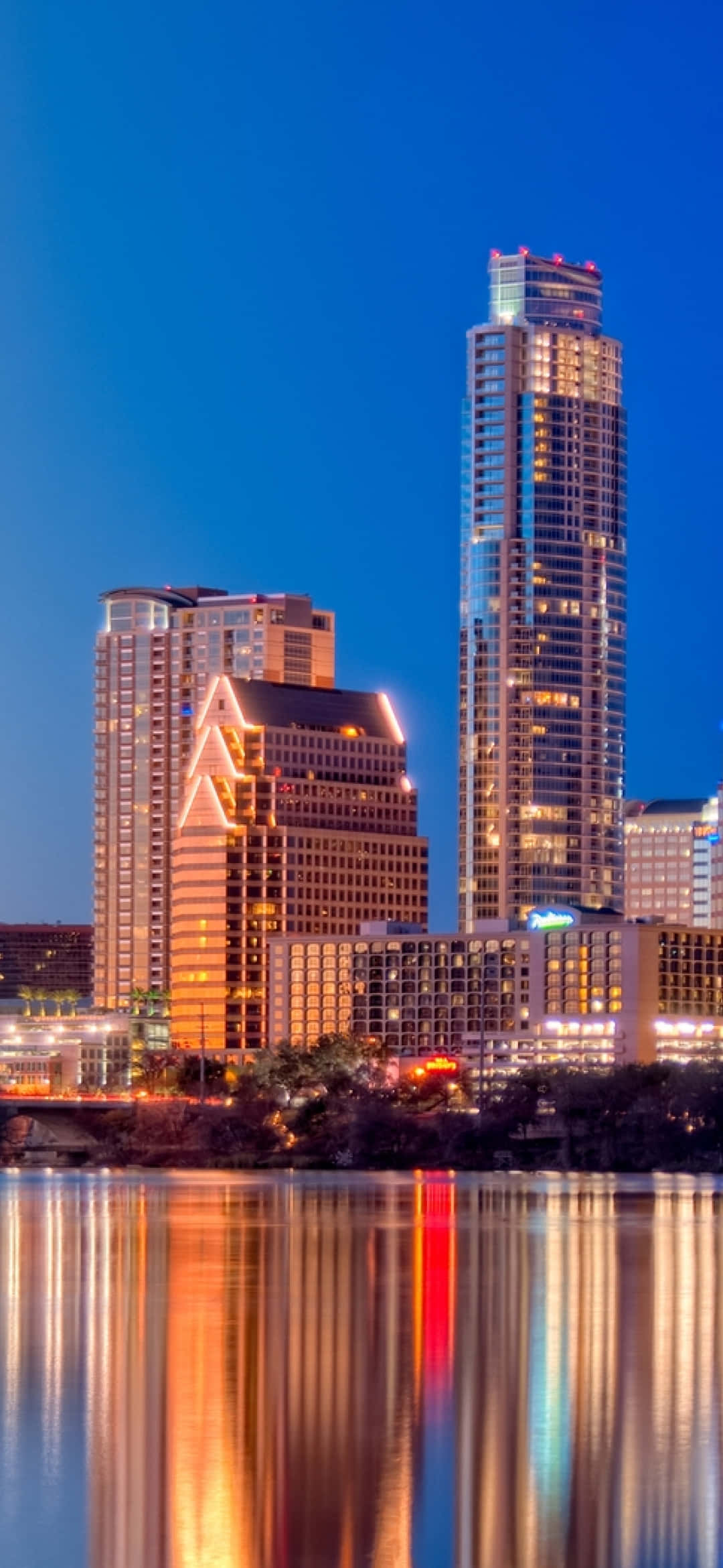 Texas iPhone 桌布 壁紙 — 高聳的城市摩天大樓在深藍色的暮色天空下散發著溫暖的光芒，它們的倒影在下方平靜的河面上閃爍生輝。
