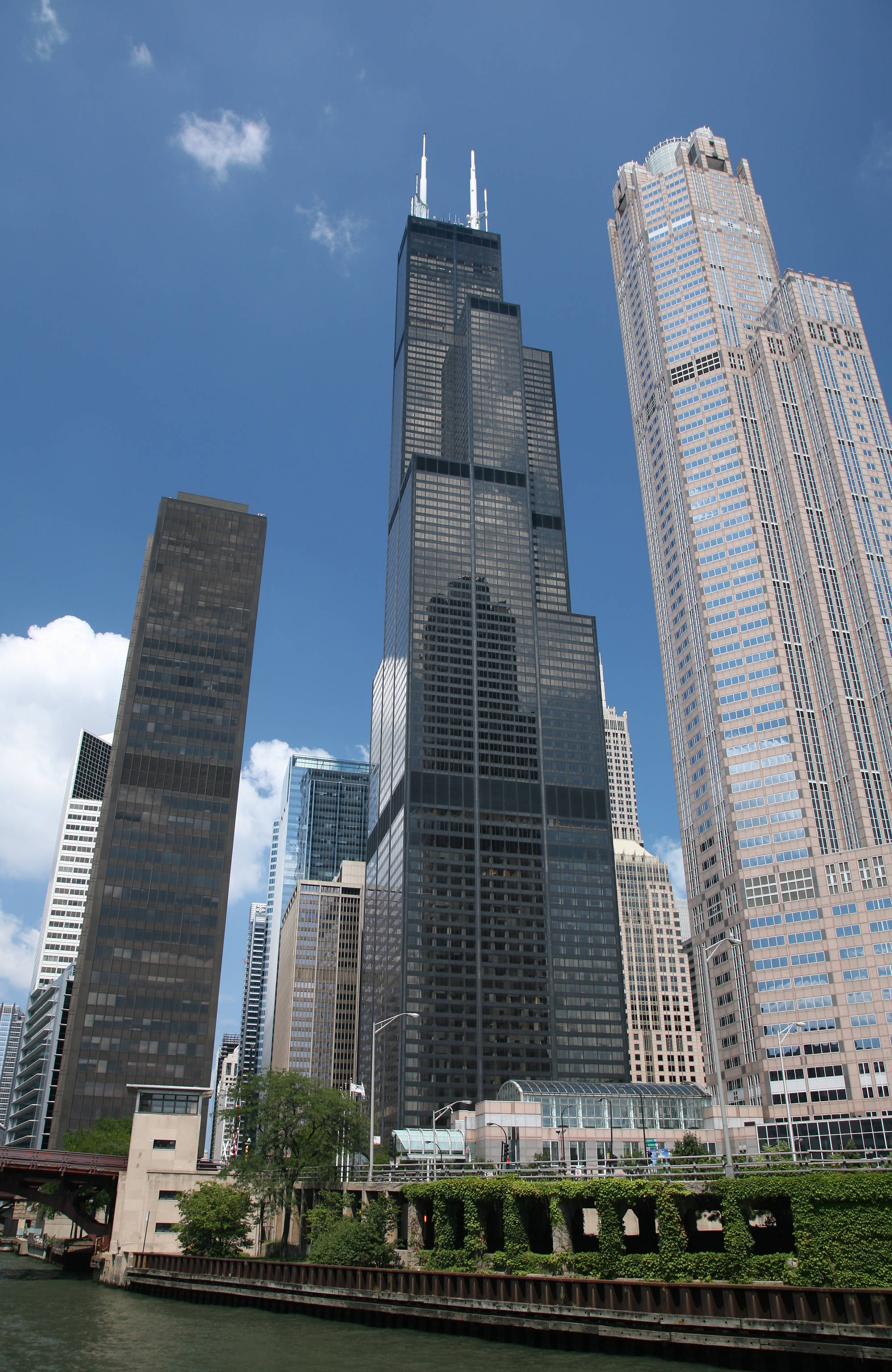 Buildings 桌布 壁紙 — 高聳的摩天大樓刺穿明亮的藍天，天空中有蓬鬆的白雲，下方是綠樹成蔭的河流。