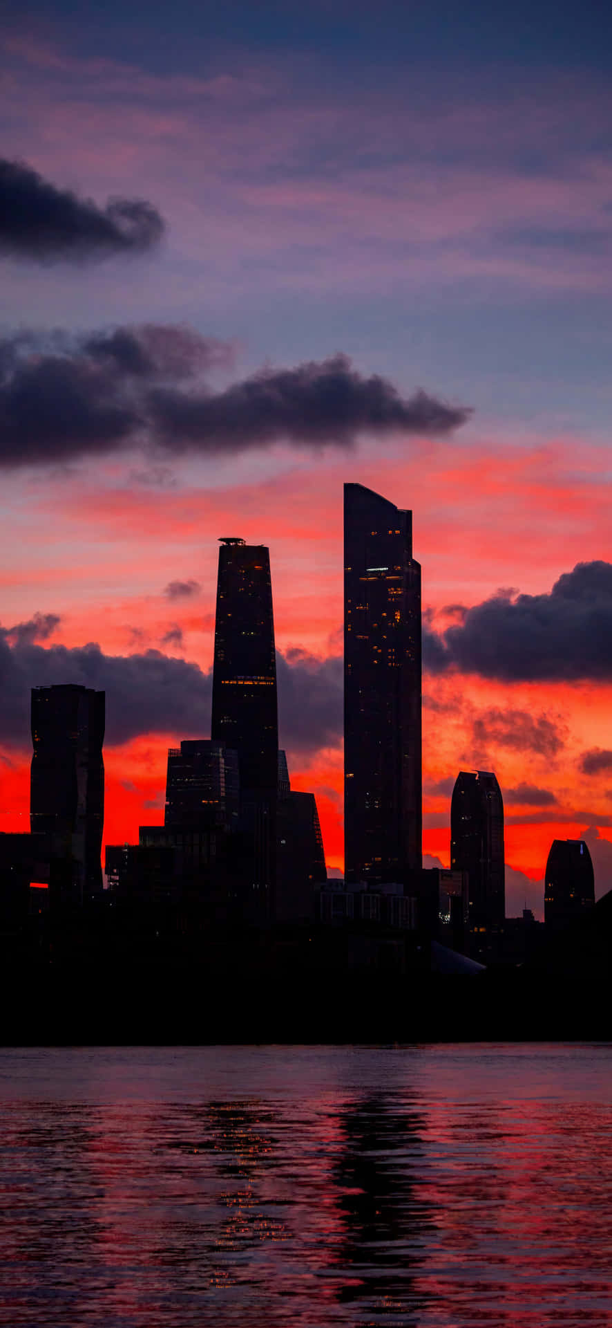 Dark city towers at sunset 4K 壁紙 — 高聳的摩天大樓在平靜的水面上映照著火紅的夕陽天空，形成黑暗的剪影。