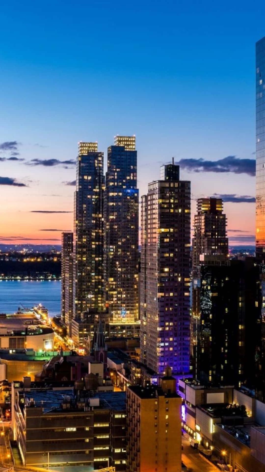 Android Ny City 桌布 壁紙 — 高聳的摩天大樓在暮色天空下閃爍著溫暖的光芒，河流映照出城市充滿活力的能量。