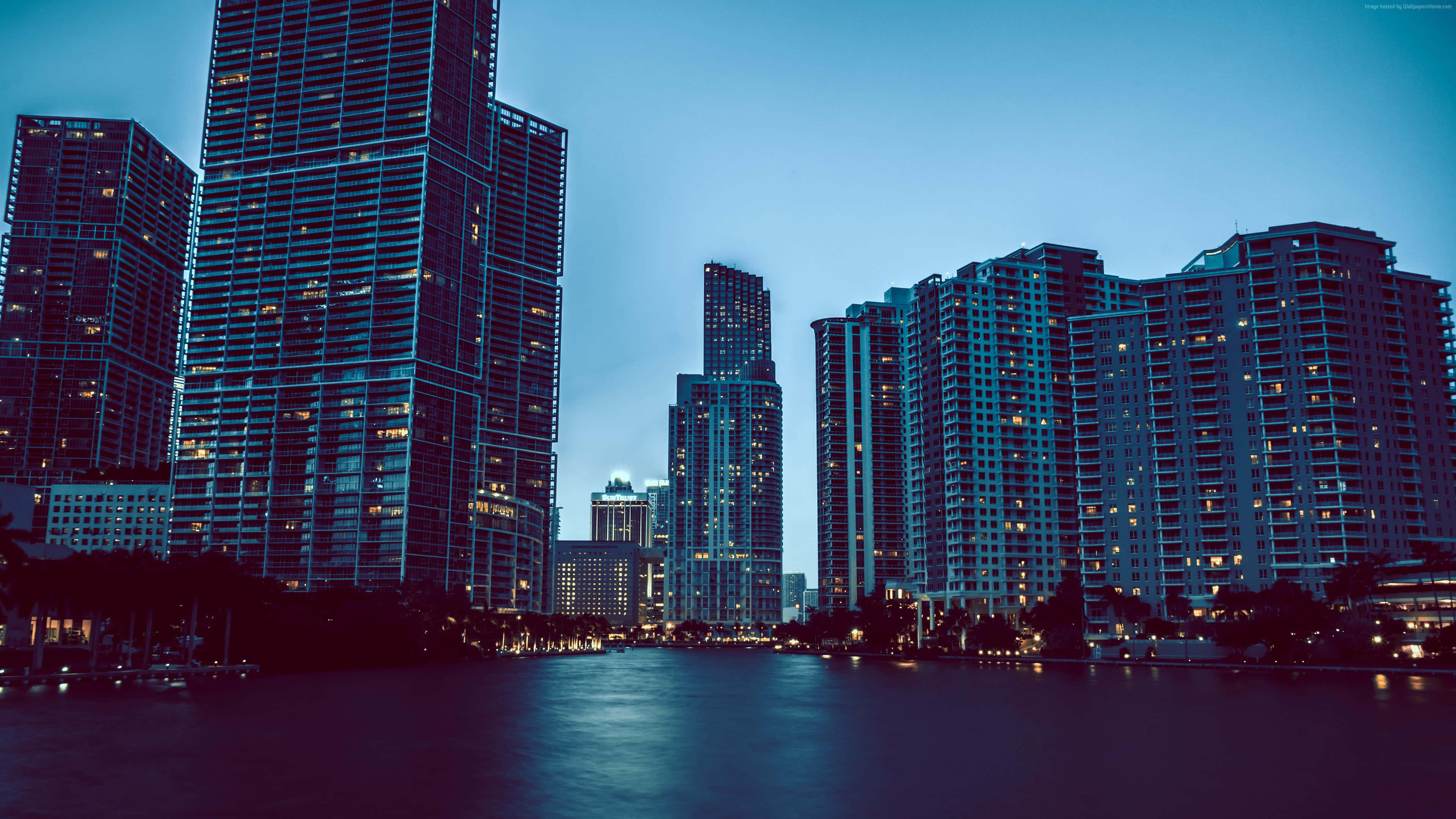 Miami 4K 桌布 壁紙 — 高聳的摩天大樓在暮色天空的映襯下散發著溫暖的光芒，倒映在平靜的水面上。