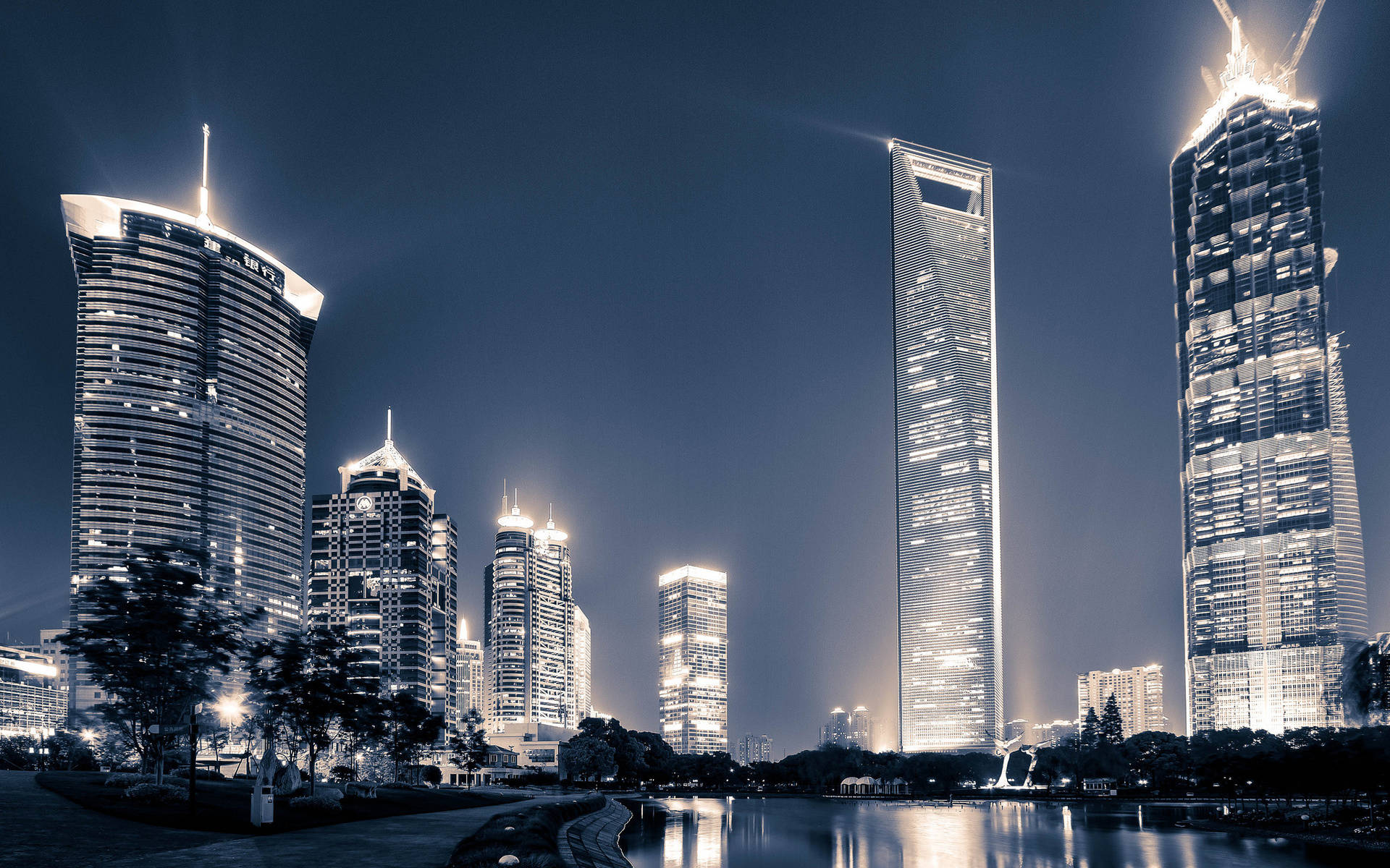 Shanghai 桌布 壁紙 — 高聳的摩天大樓在深藍色的夜空中閃耀，它們的燈光倒映在下方平靜的海濱上。