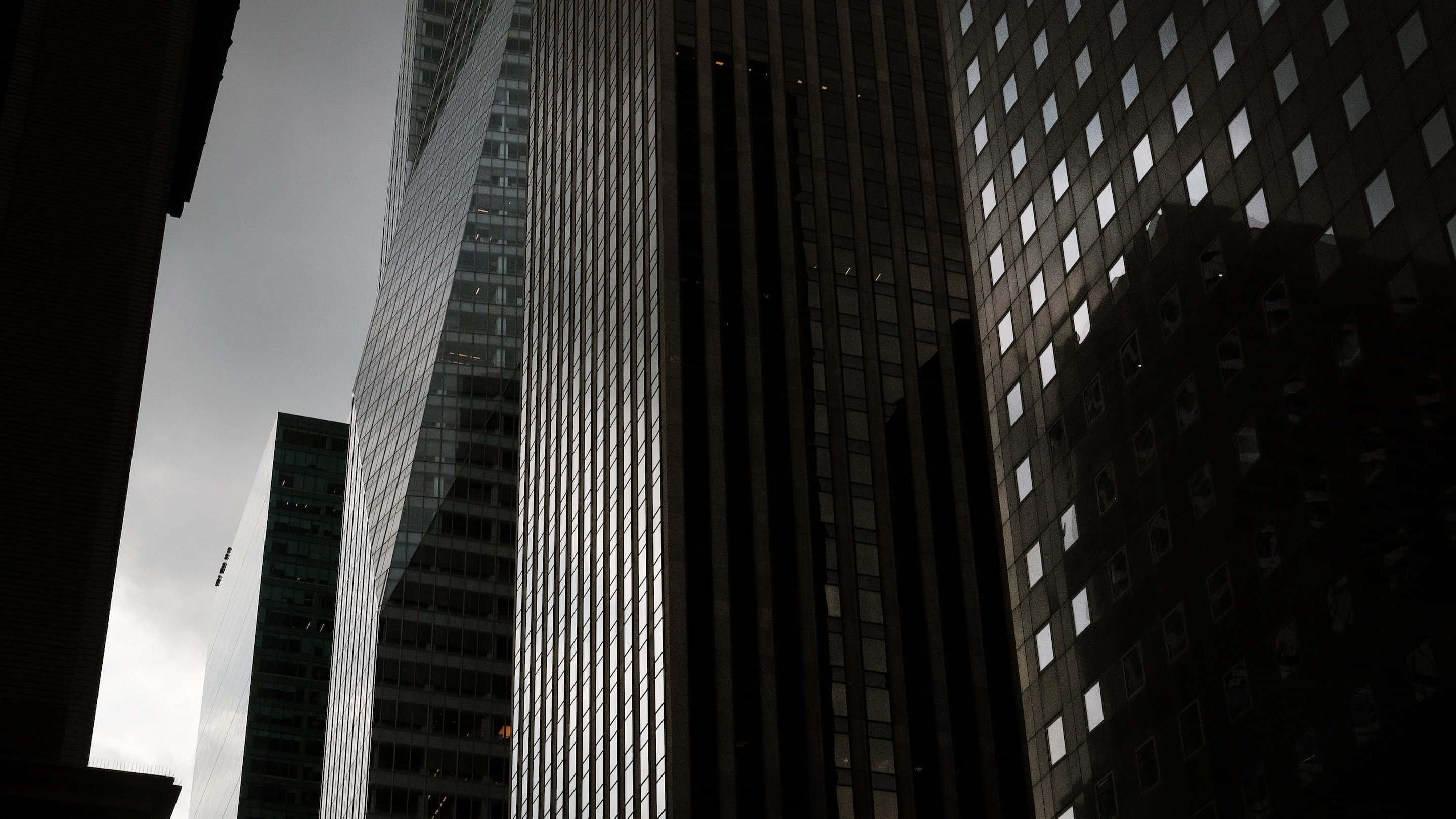 Buildings 桌布 壁紙 — 高聳的摩天大樓在陰沉天空下若隱若現，帶有鮮明的垂直線條和捕捉微弱光線的反射玻璃窗。