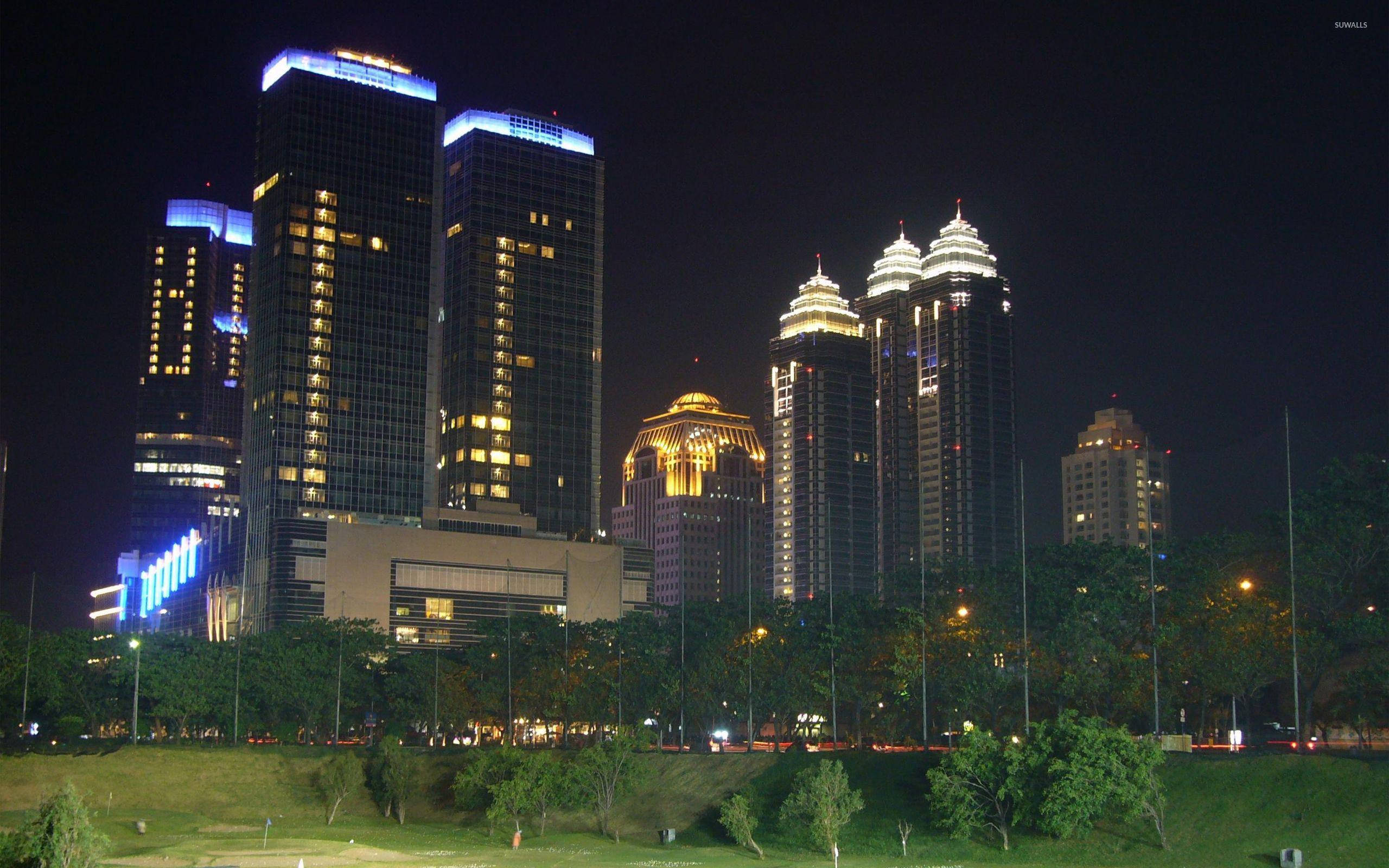 Jakarta 桌布 壁紙 — 高聳的摩天大樓在黑暗的夜空中閃爍著藍色和金色的燈光，下方是綠色的公園，有樹木和路燈。