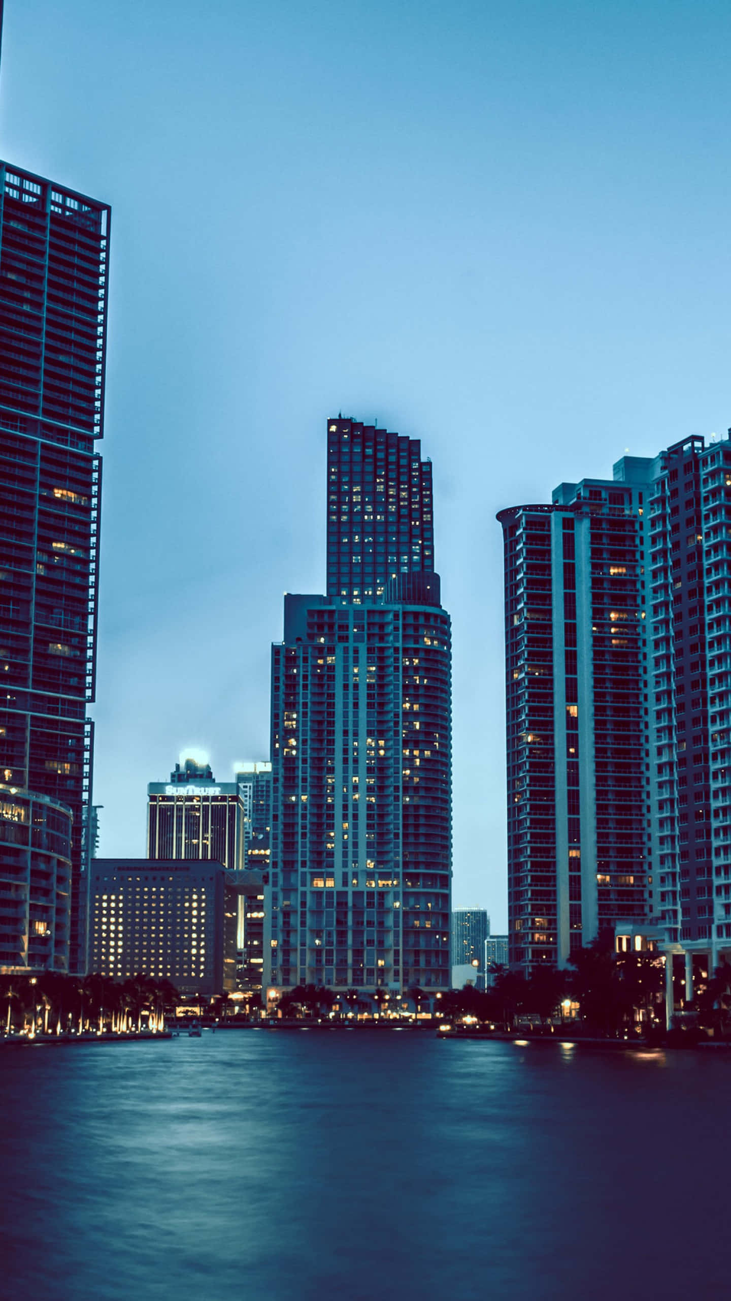 Miami 4K 桌布 壁紙 — 高聳的摩天大樓閃爍著窗戶燈光，排列在寧靜的海濱，天空呈現暮光藍色。