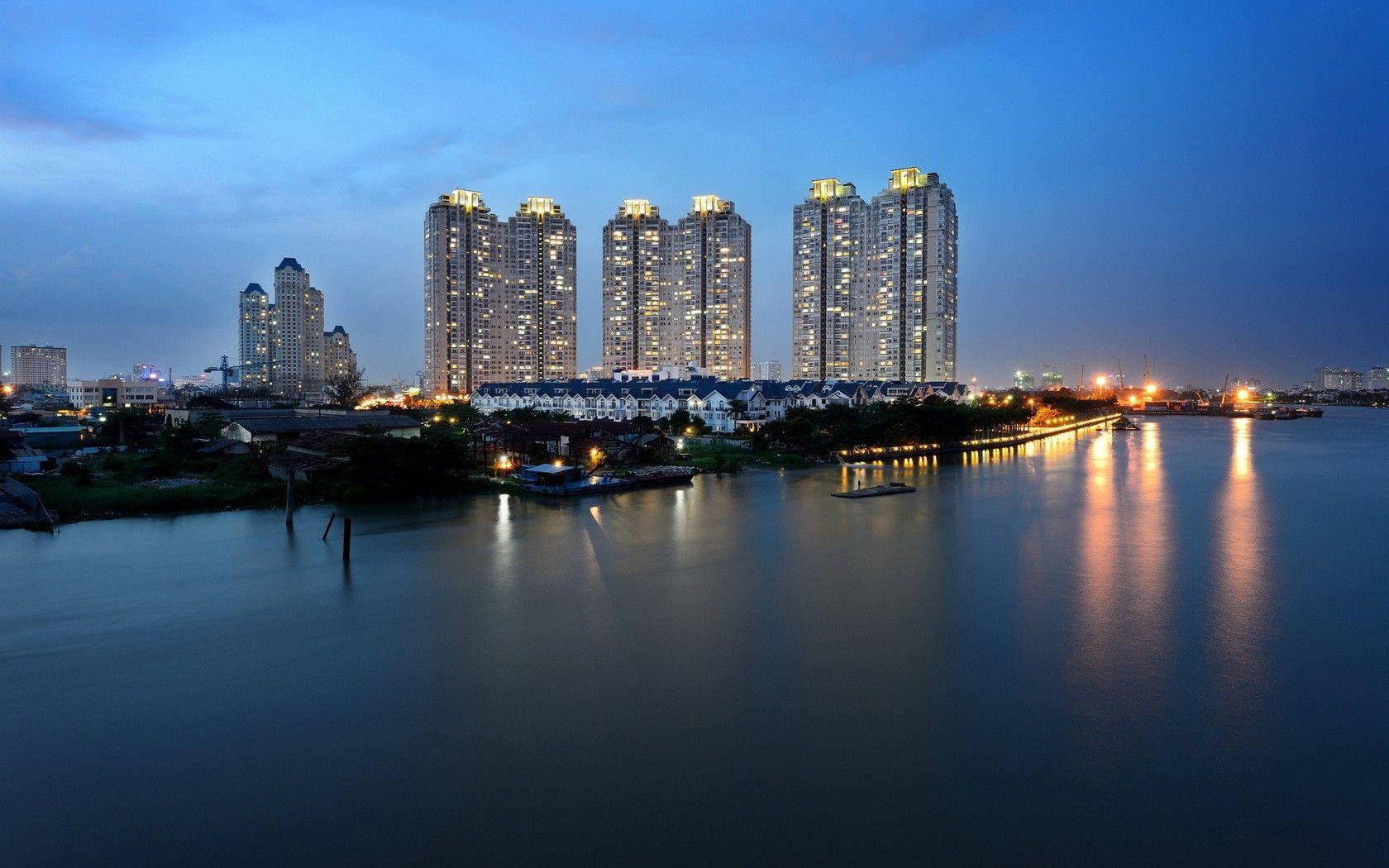 Ho Chi Minh City 桌布 壁紙 — 高聳的燈火通明摩天大樓在暮色中倒映在平靜的水面上，遠處有橋樑閃爍著光芒。