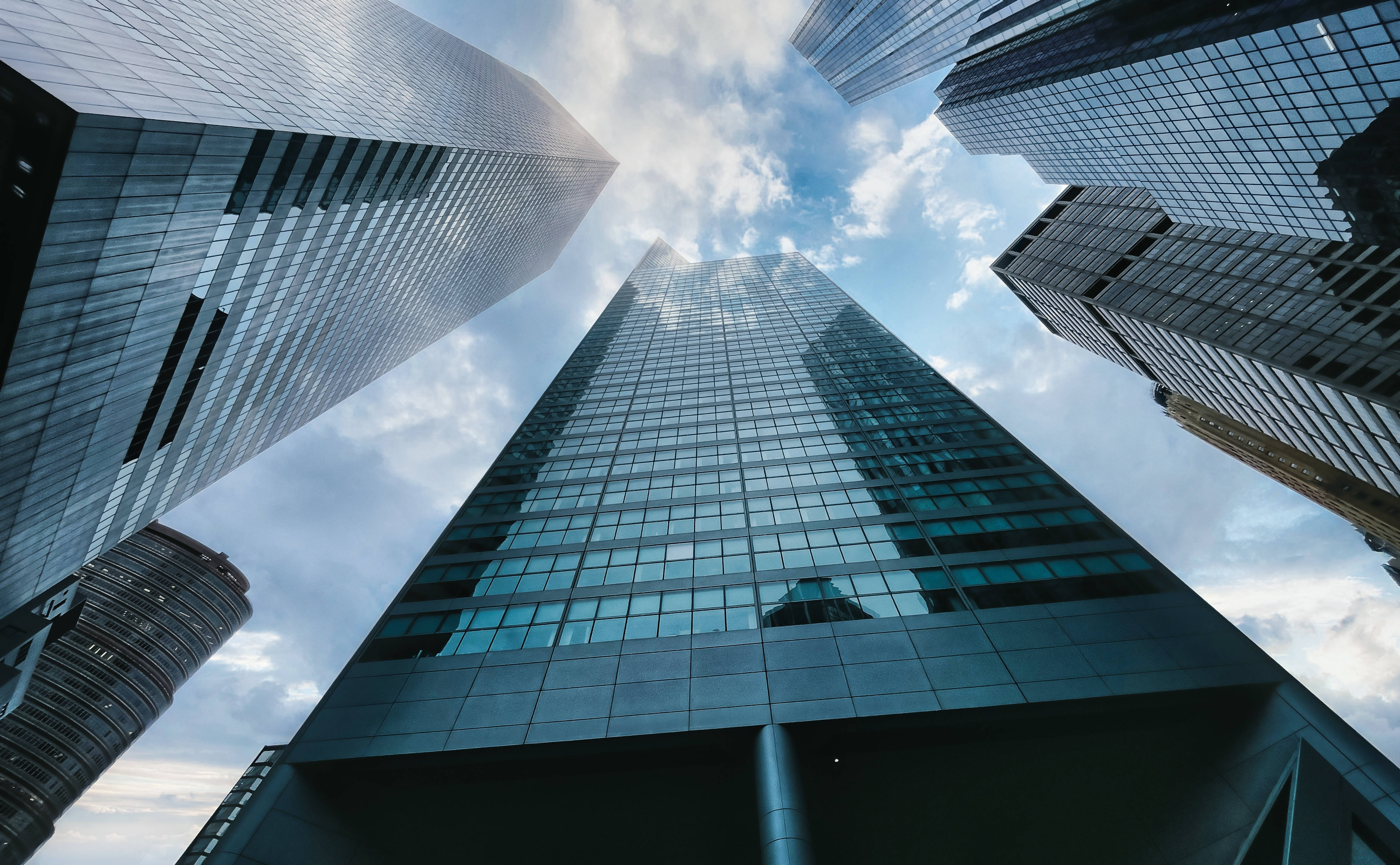 Skyscraper 桌布 壁紙 — 高聳的玻璃摩天大樓刺穿明亮藍天，散佈著雲朵，戲劇性的低角度城市拍攝。