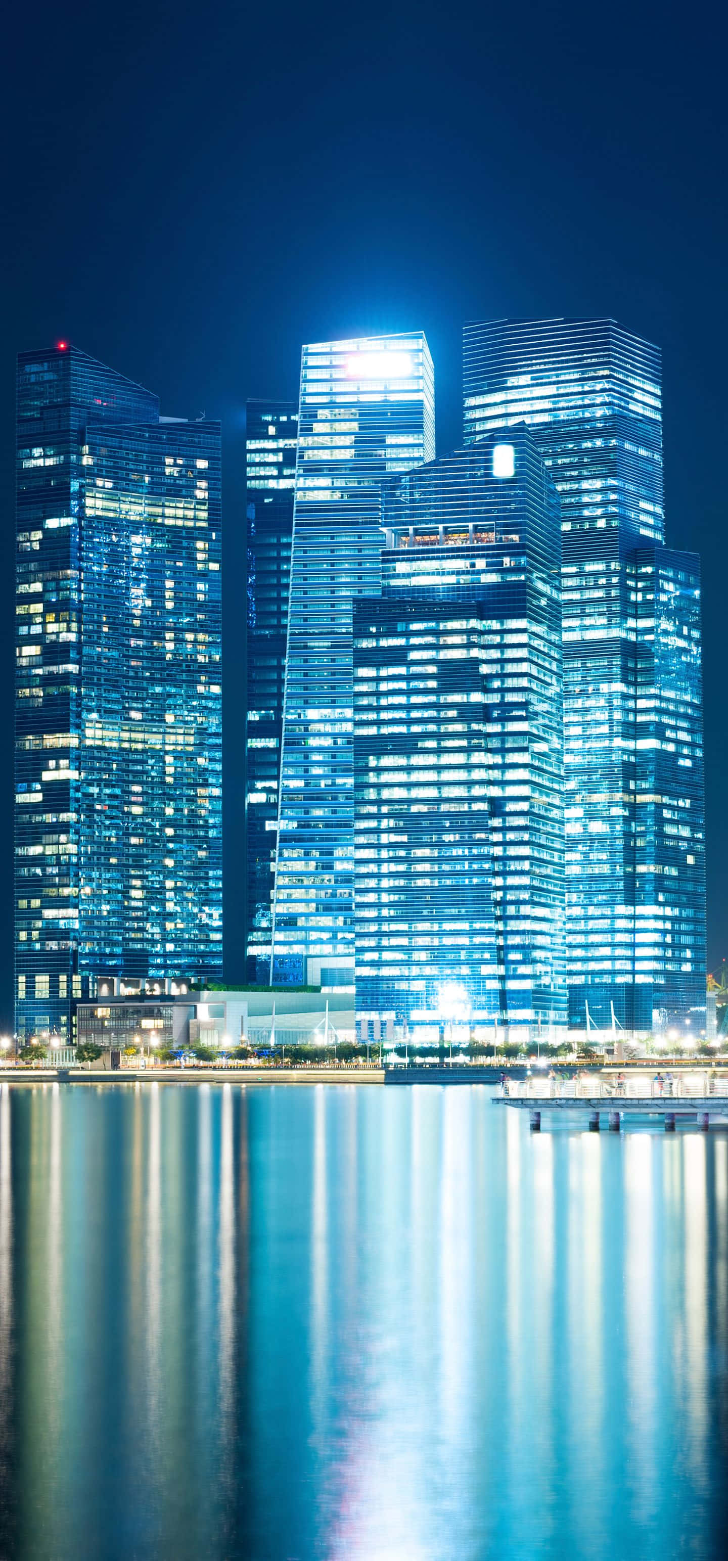 1440x3088 桌布 壁紙 — 高聳的玻璃摩天大樓在黑暗的夜空中發出藍光，它們的倒影在平靜的水面上閃爍。