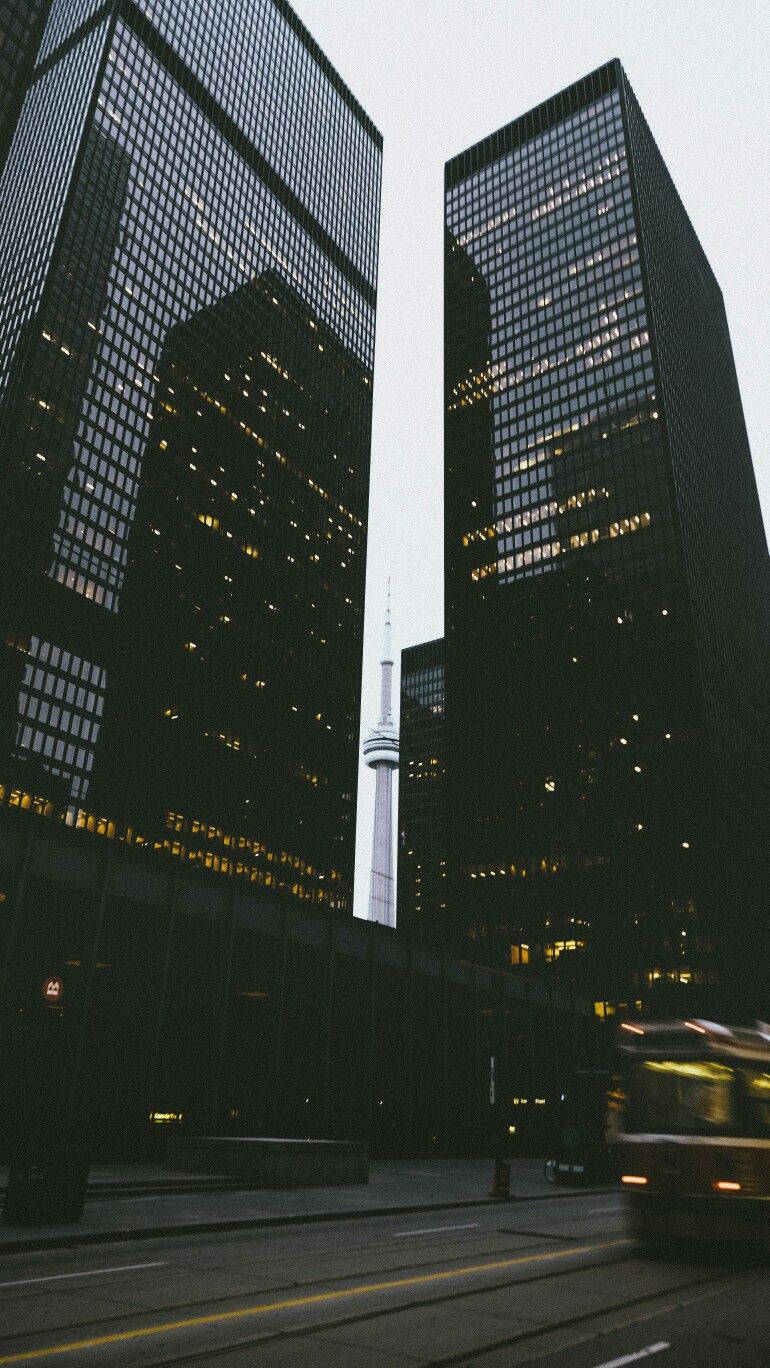 Buildings 桌布 壁紙 — 高聳的玻璃摩天大樓框住一座纖細的塔樓，一輛模糊的巴士在黃昏的城市街道上疾馳而過。