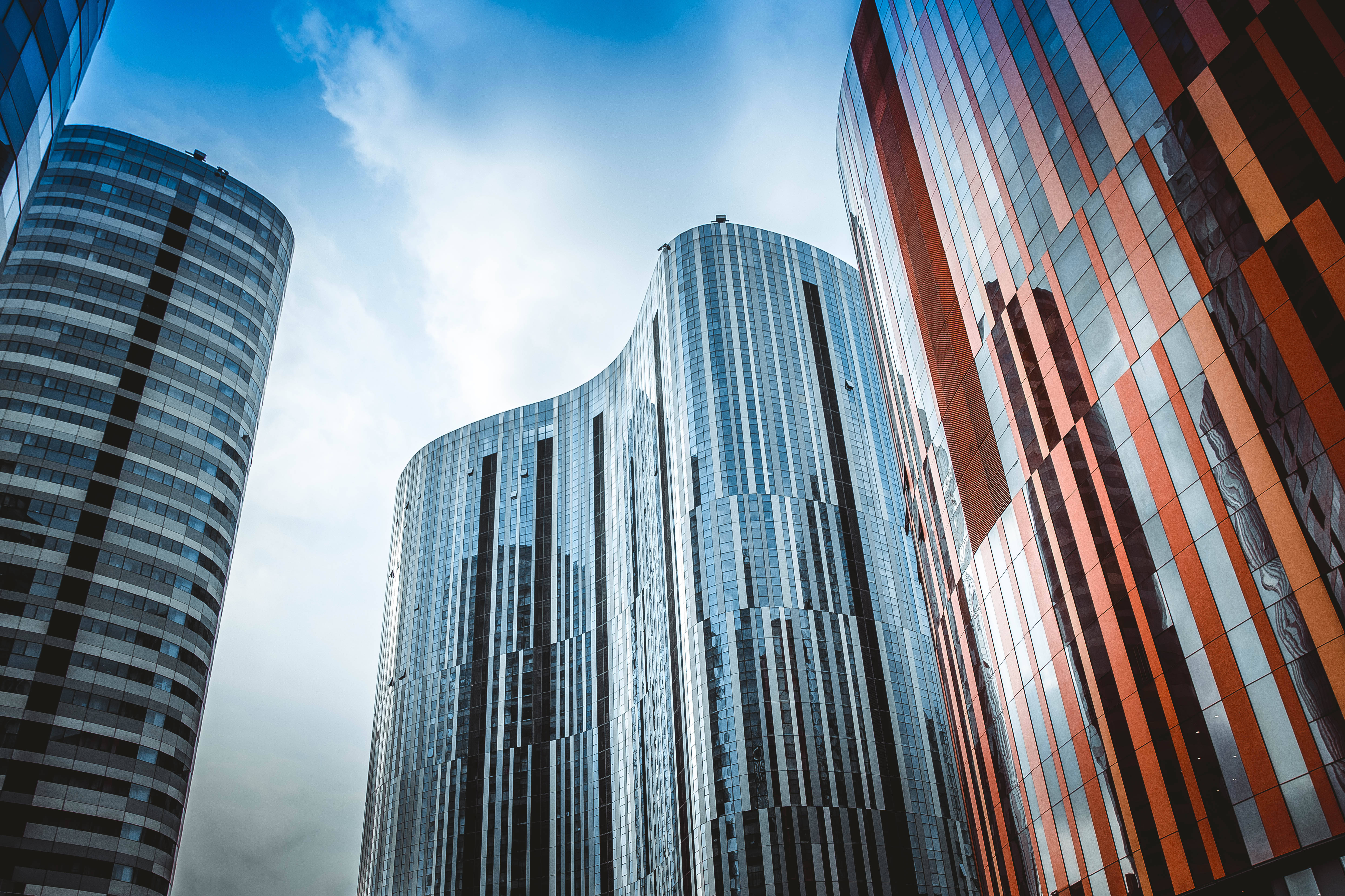 Skyscraper 桌布 壁紙 — 高聳的現代摩天大樓，帶有弧形玻璃外牆，在戲劇性的低角度城市拍攝中反射藍天和白雲。