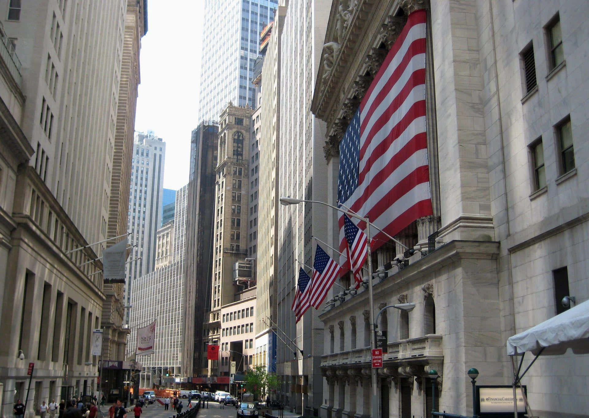 Wall Street 桌布 壁紙 — 高聳的石建築物排列在繁忙的城市街道上，大型美國國旗從歷史悠久的金融機構飄揚。