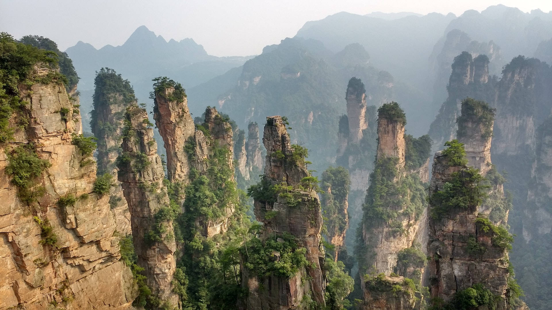 Mountain Landscape 桌布 壁紙 — 高聳的砂岩柱從薄霧籠罩的山谷中升起，位於鬱鬱蔥蔥的綠色山脈中，沐浴在柔和的白日光下。