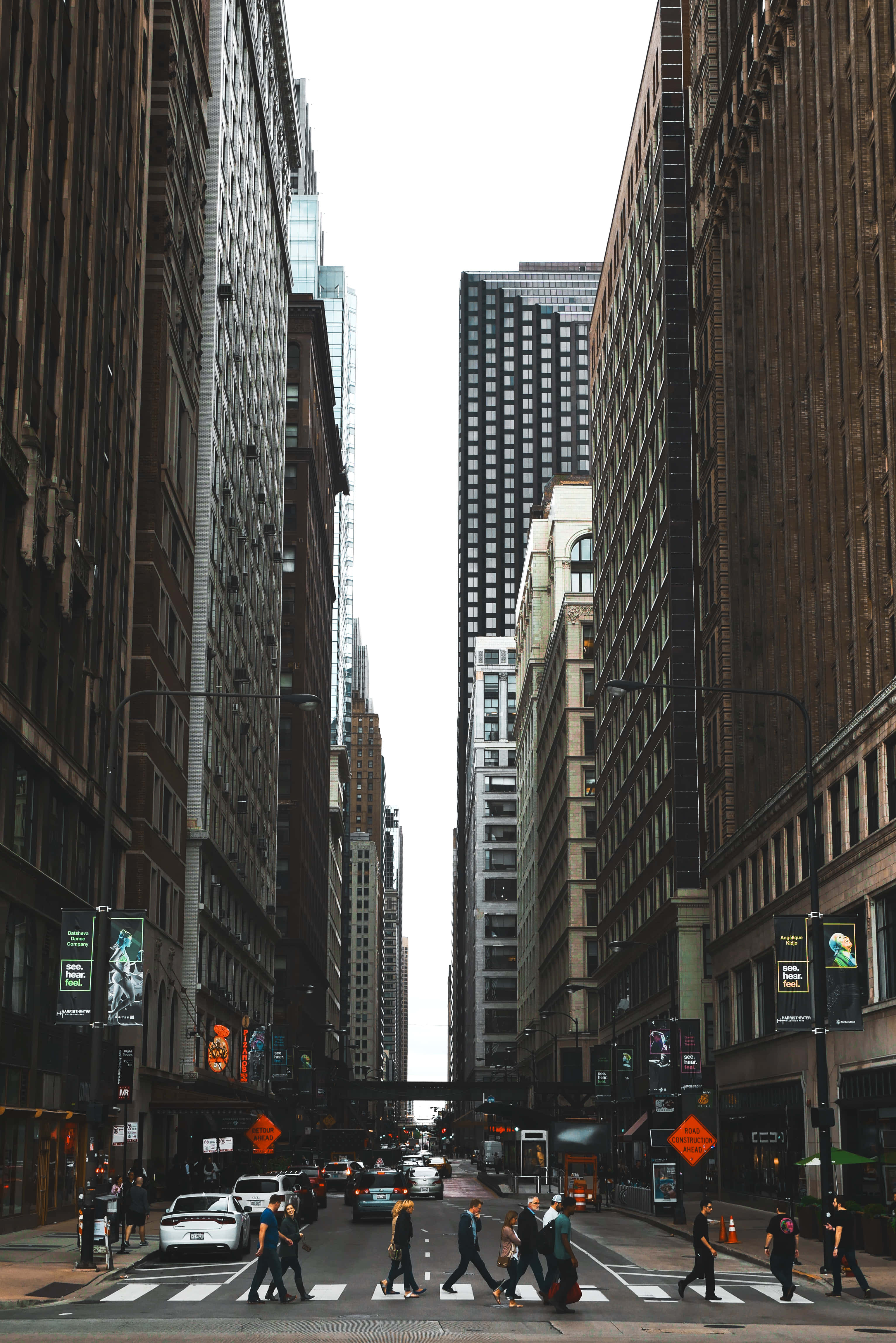 Wall Street 桌布 壁紙 — 高聳的磚和玻璃摩天大樓框住一條繁忙的城市街道，行人在陰天的人行橫道上過馬路。