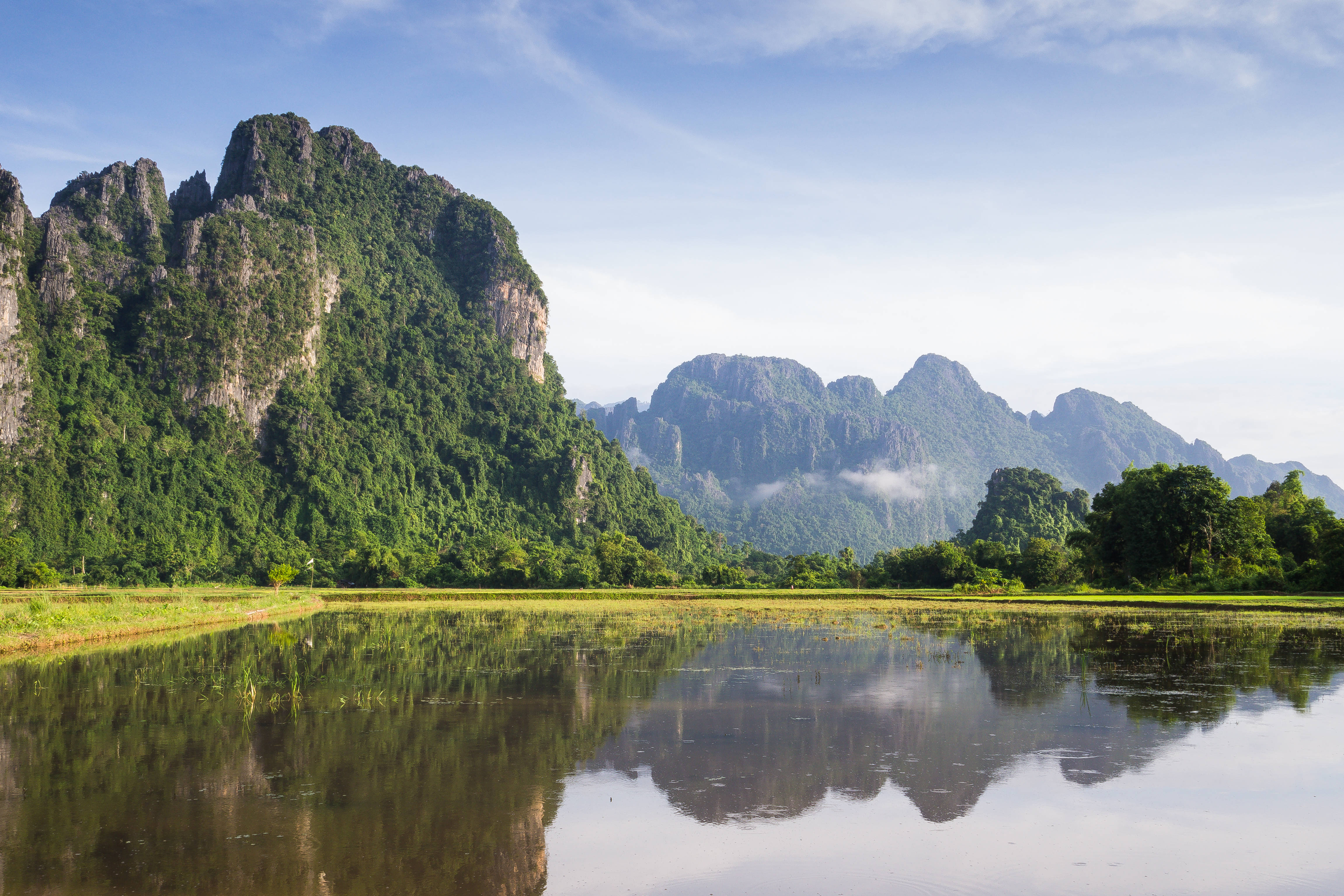 Laos 桌布 壁紙 — 高聳的綠色喀斯特山脈在明亮的藍天下，倒映在平靜的水稻田中，呈現出雄偉的輪廓。