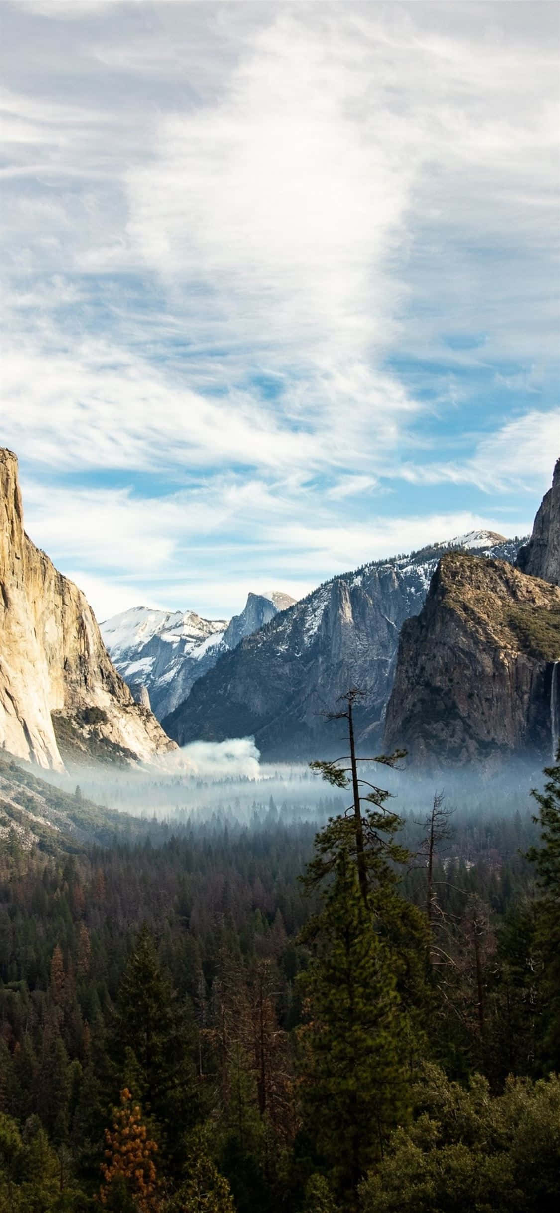 iPhone X California 桌布 壁紙 — 高聳的花崗岩懸崖和白雪覆蓋的山峰聳立在一個充滿茂密常綠森林的霧氣山谷之上，天空部分多雲。